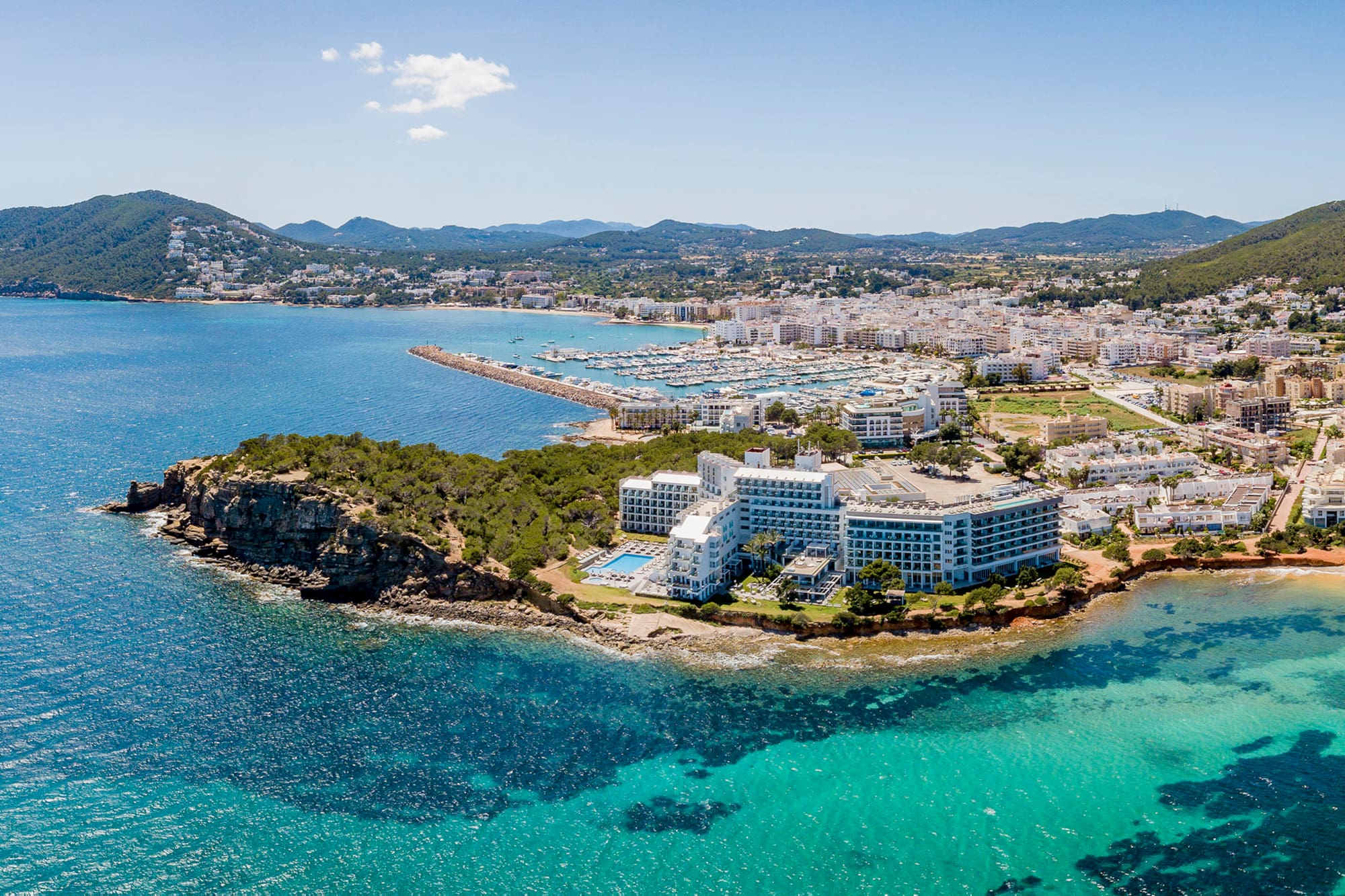 Ibiza coastal town aerial, hotels, marina, clear turquoise sea, green hills.