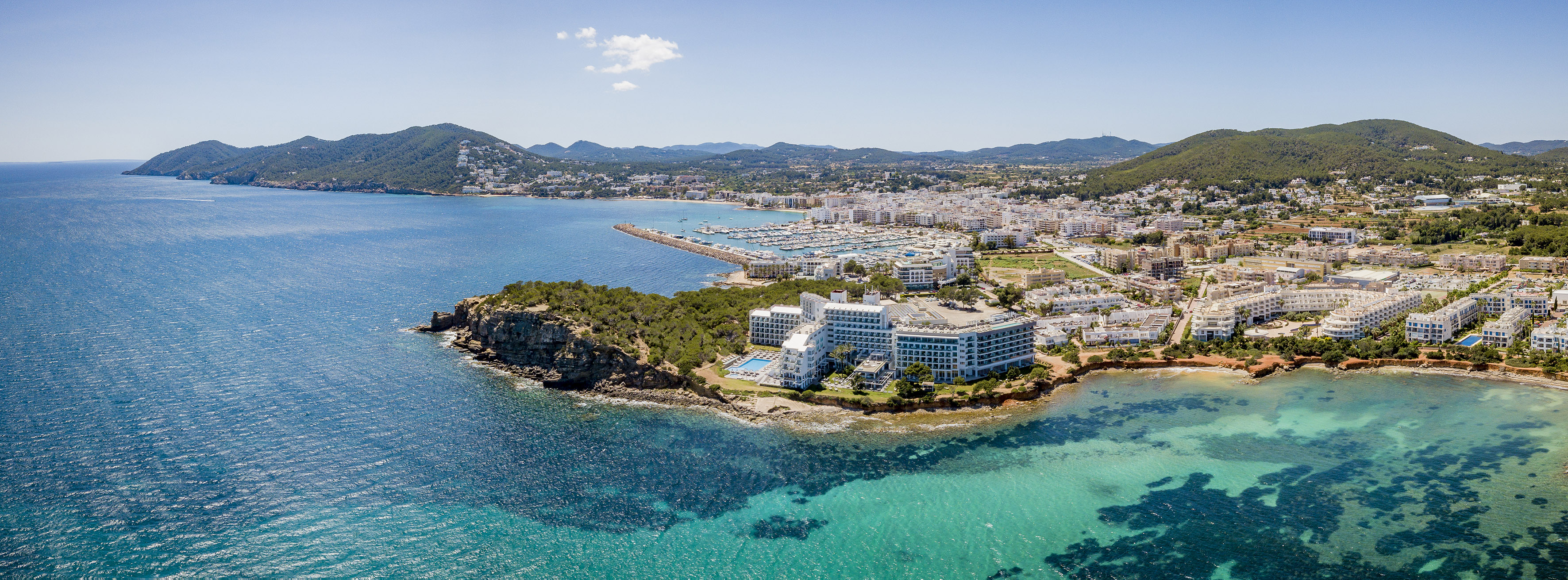Ibiza coastal town aerial, hotels, marina, clear turquoise sea, green hills.