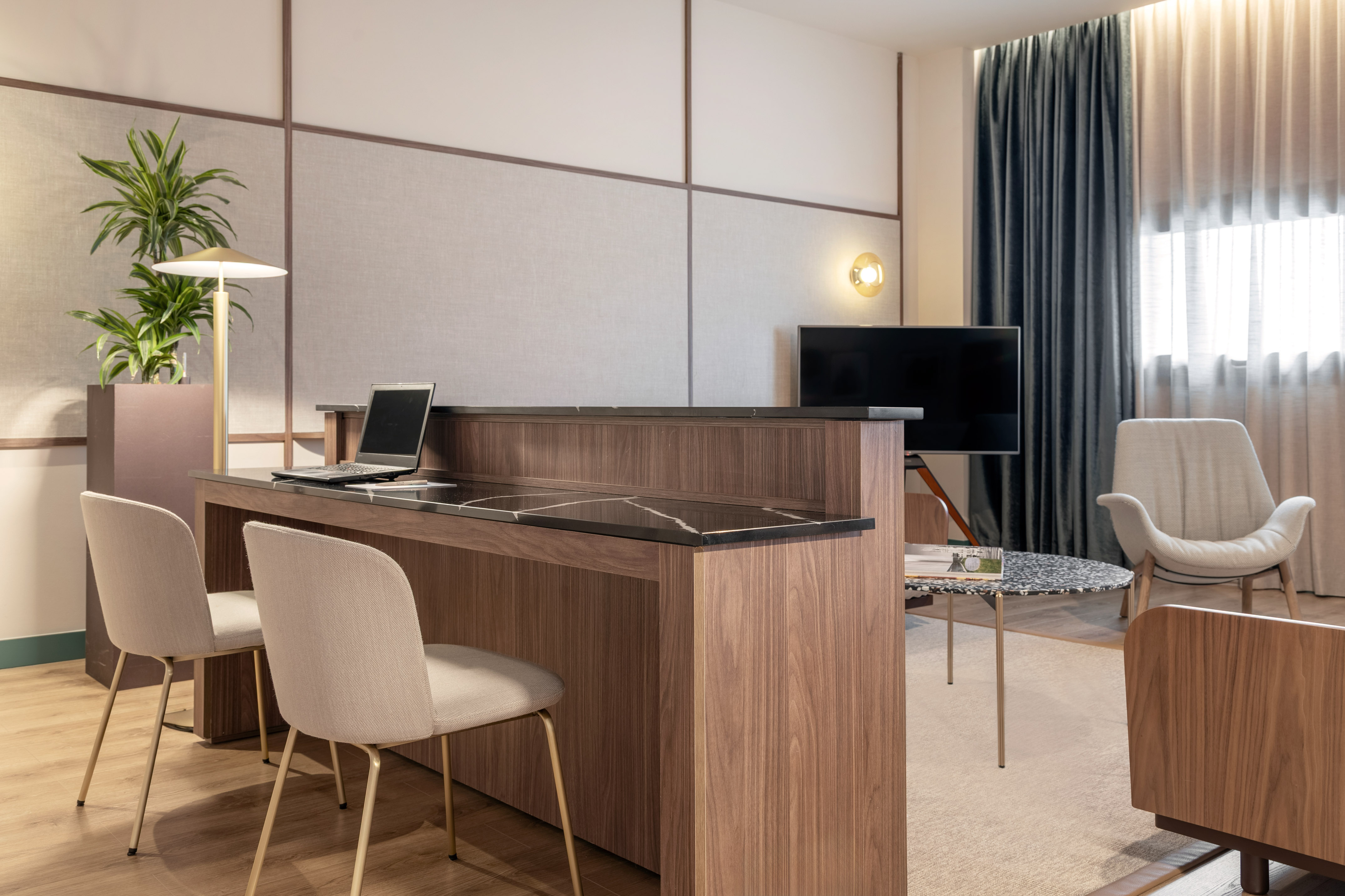 Modern hotel suite with wood and marble work desk, laptop, chairs, sitting area.