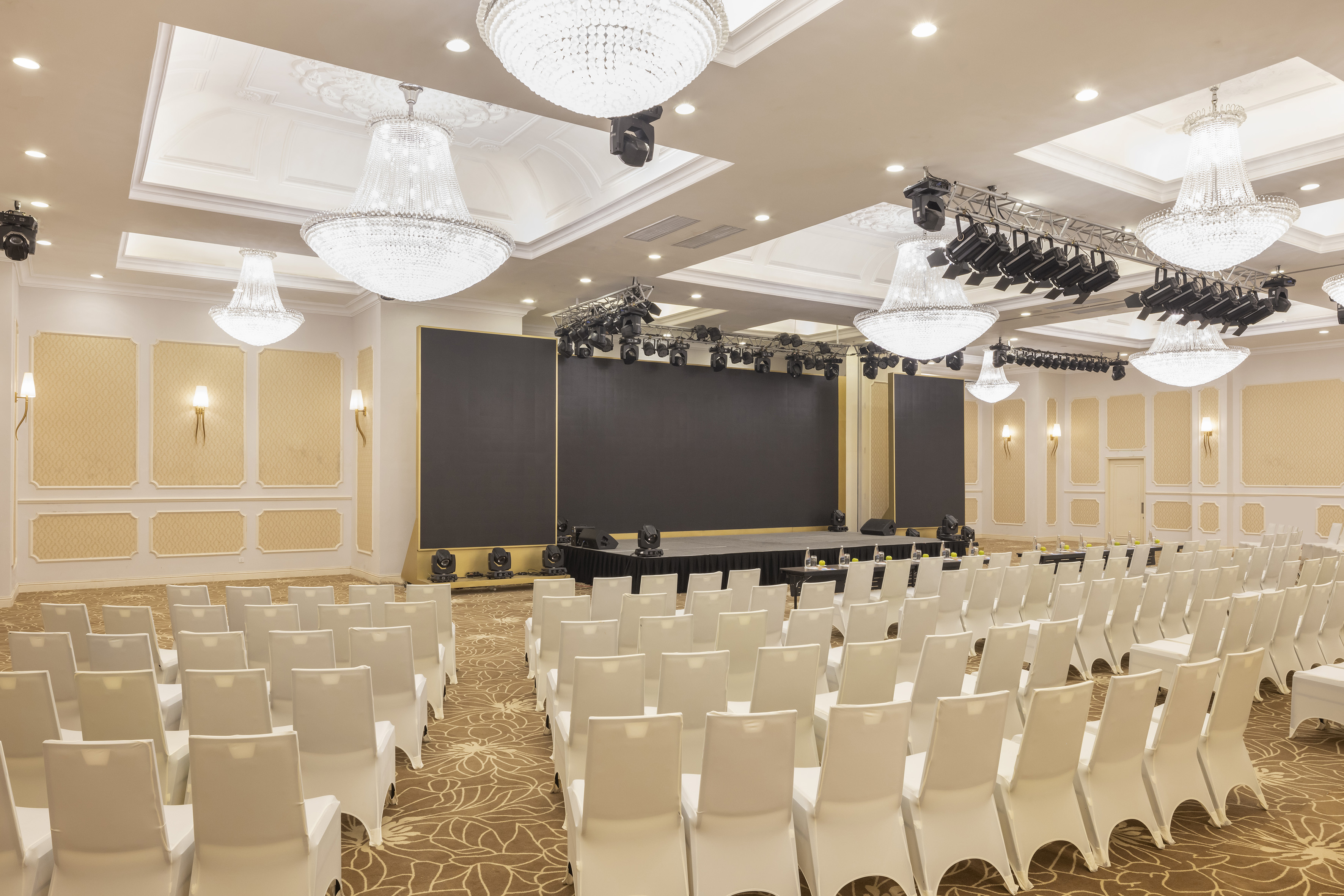 Empty meeting room with stage, LED screen, white chairs, and chandeliers.