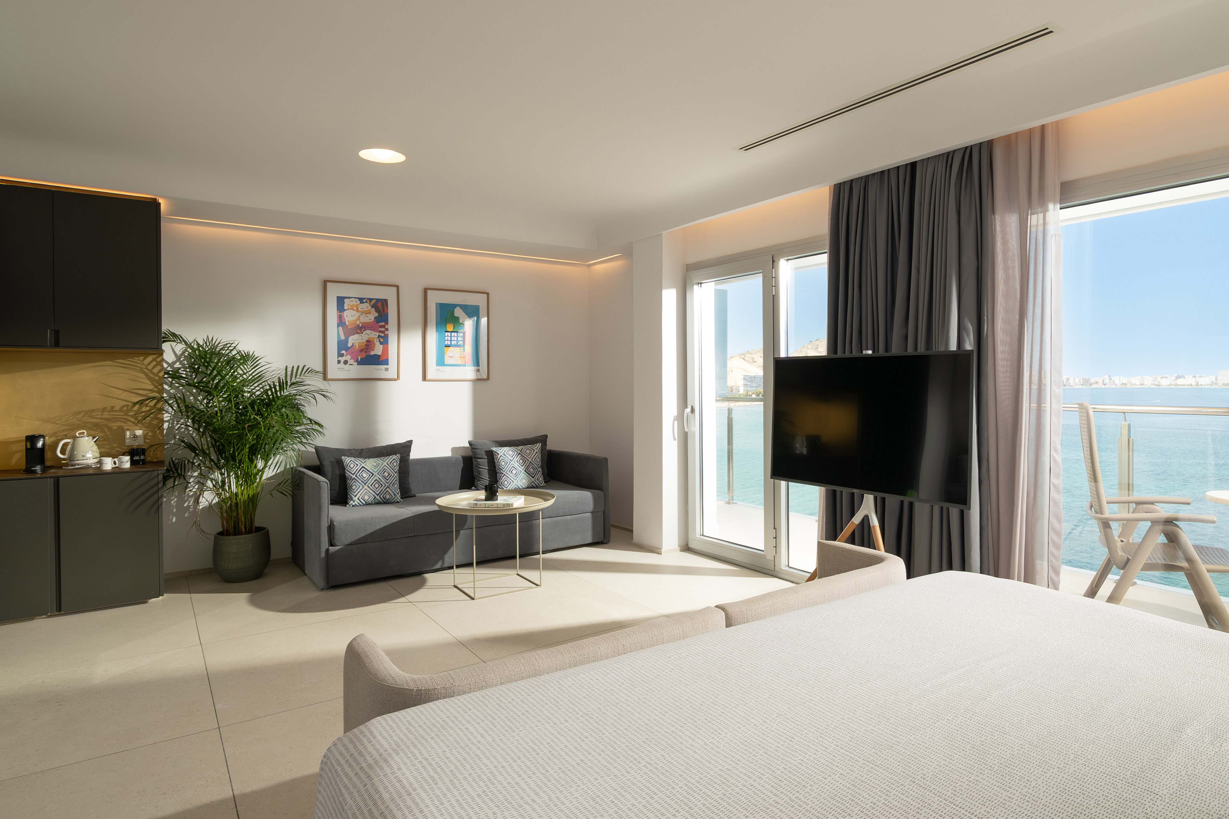 Modern hotel suite with sea view balcony, bed, sofa, TV, and contemporary decor.