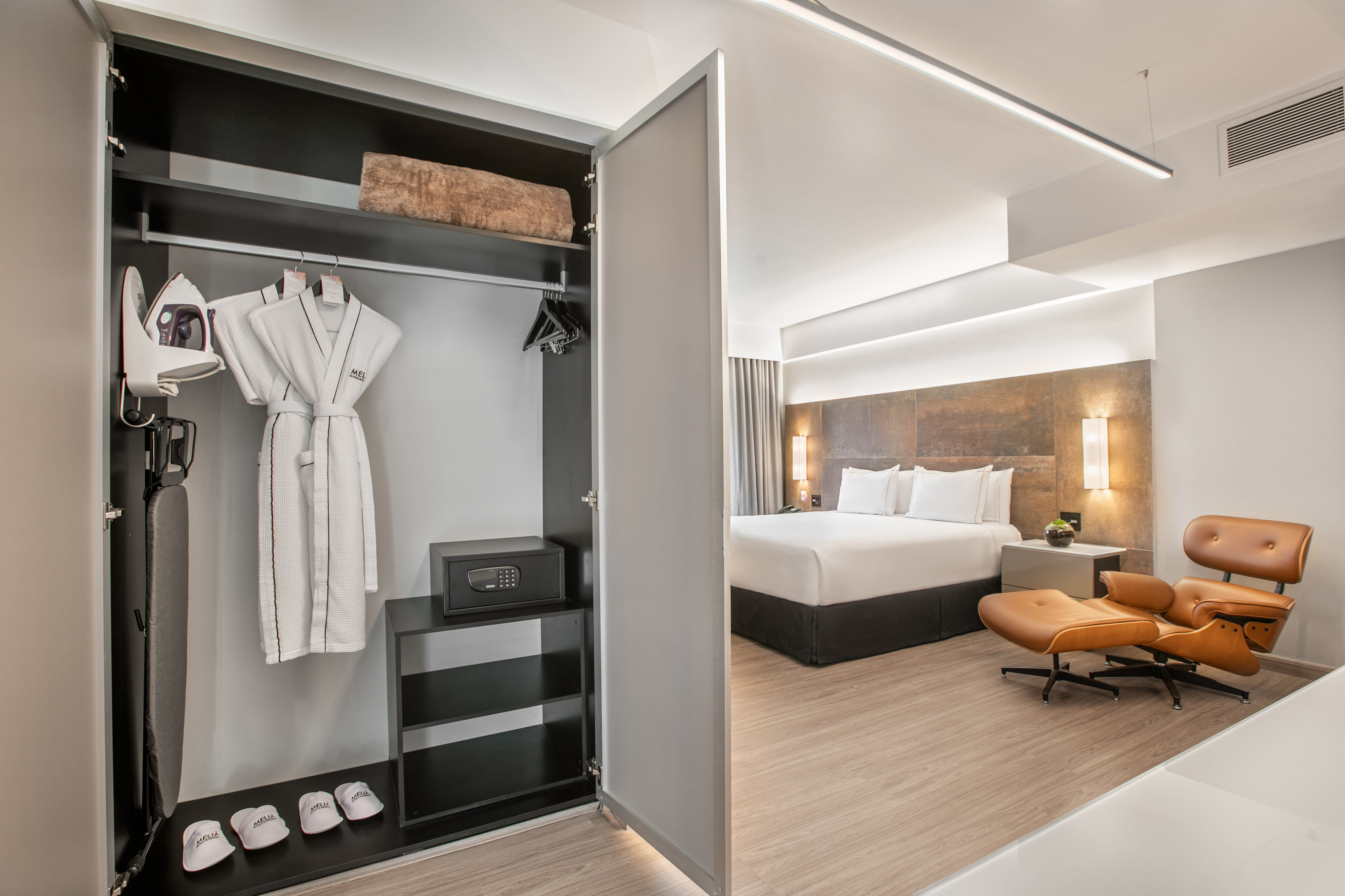 Hotel room with open closet (robes, iron, safe) and bed, lounge chair.