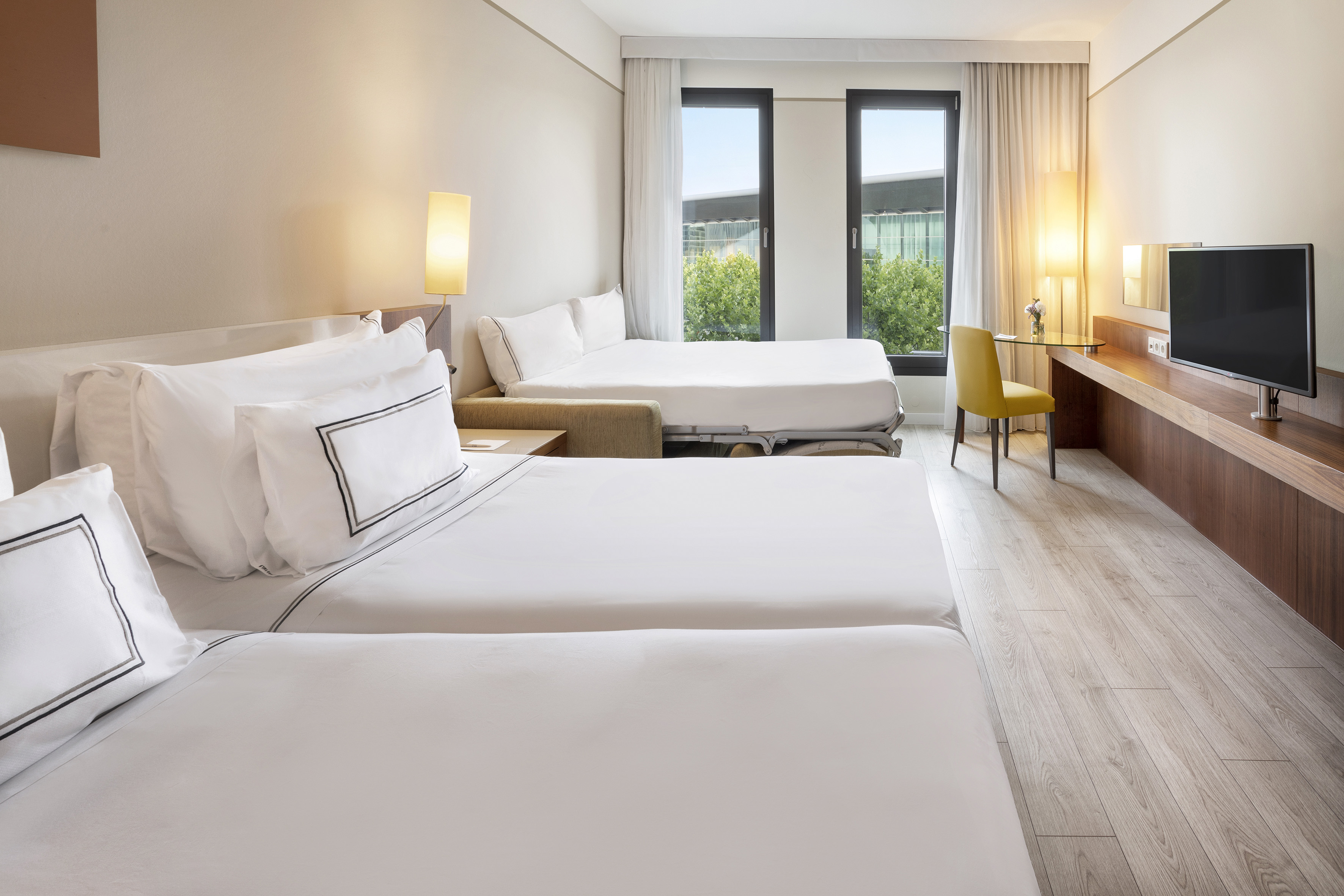 Modern hotel family room with twin beds, sofa bed, desk, and windows.