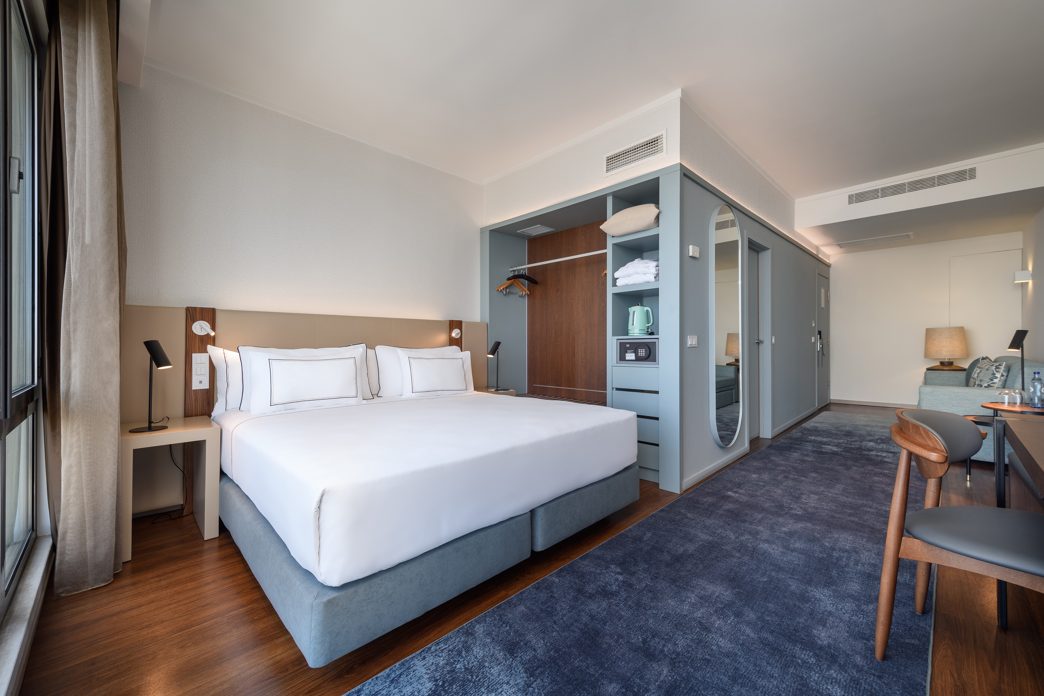 Modern hotel room with king bed, blue rug, built-in wardrobe, and desk area.