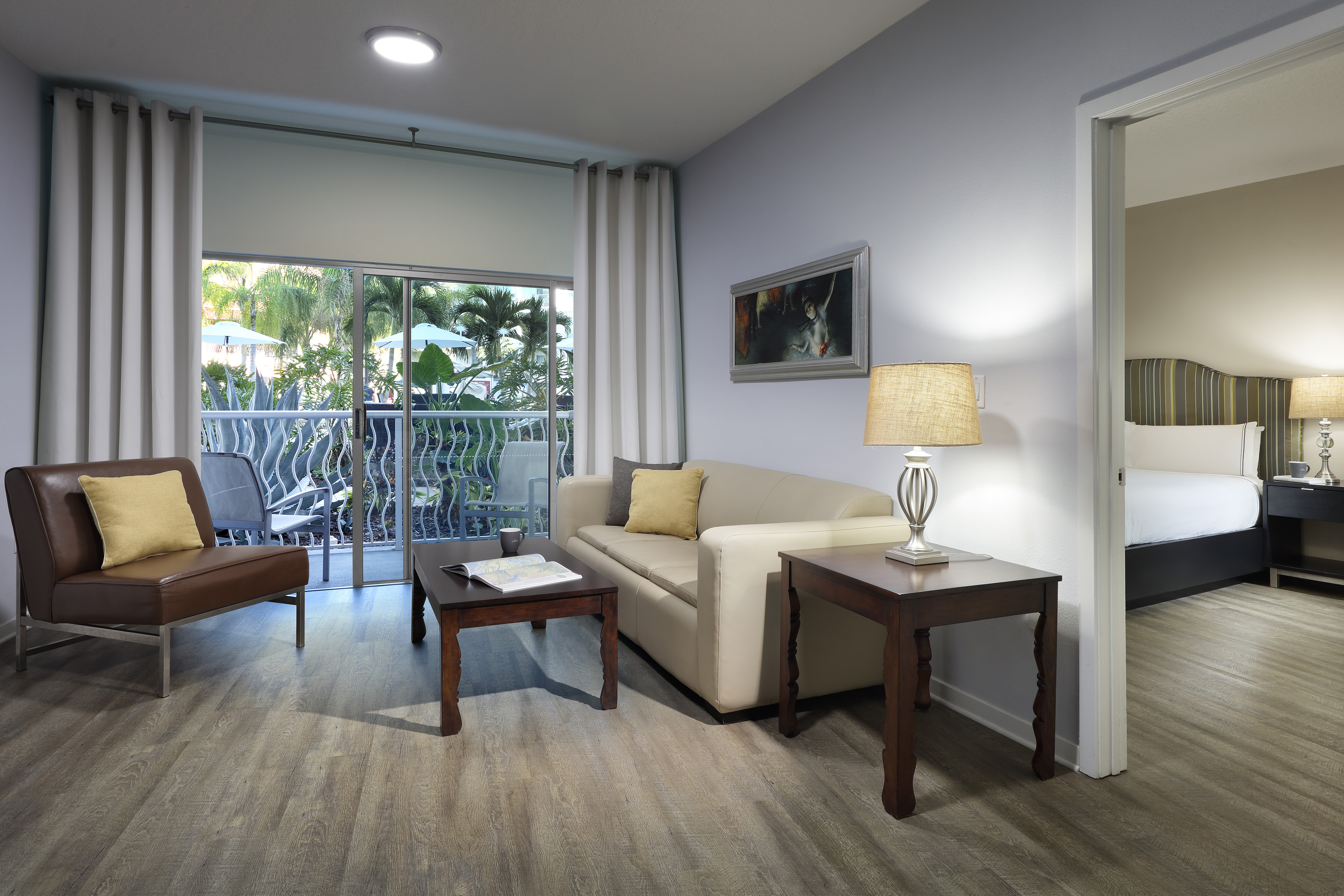 Hotel suite living area with sofa, tropical balcony view, and connected bedroom.