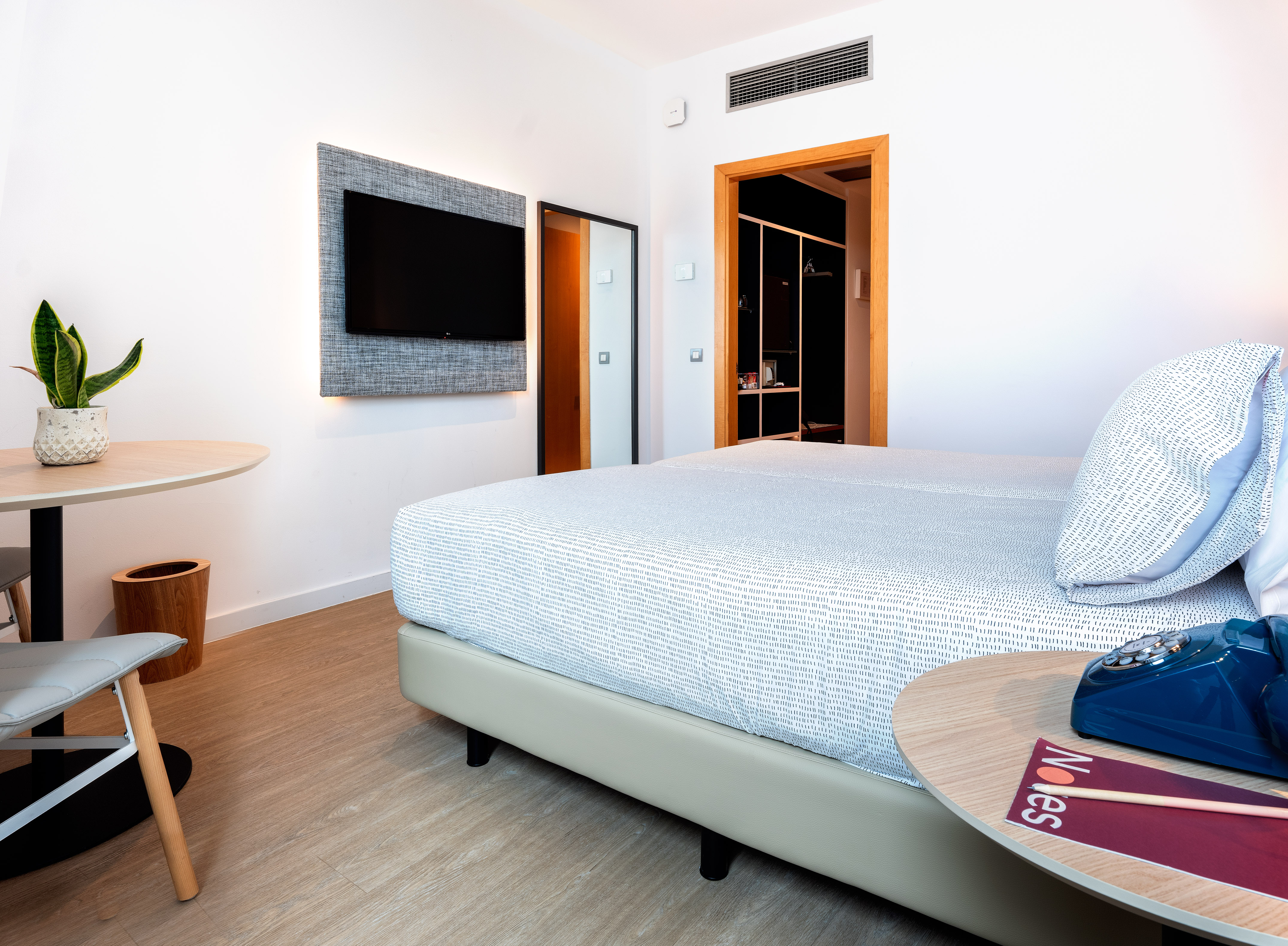 Modern hotel bedroom with white bed, wall TV, plant, and wooden floor.