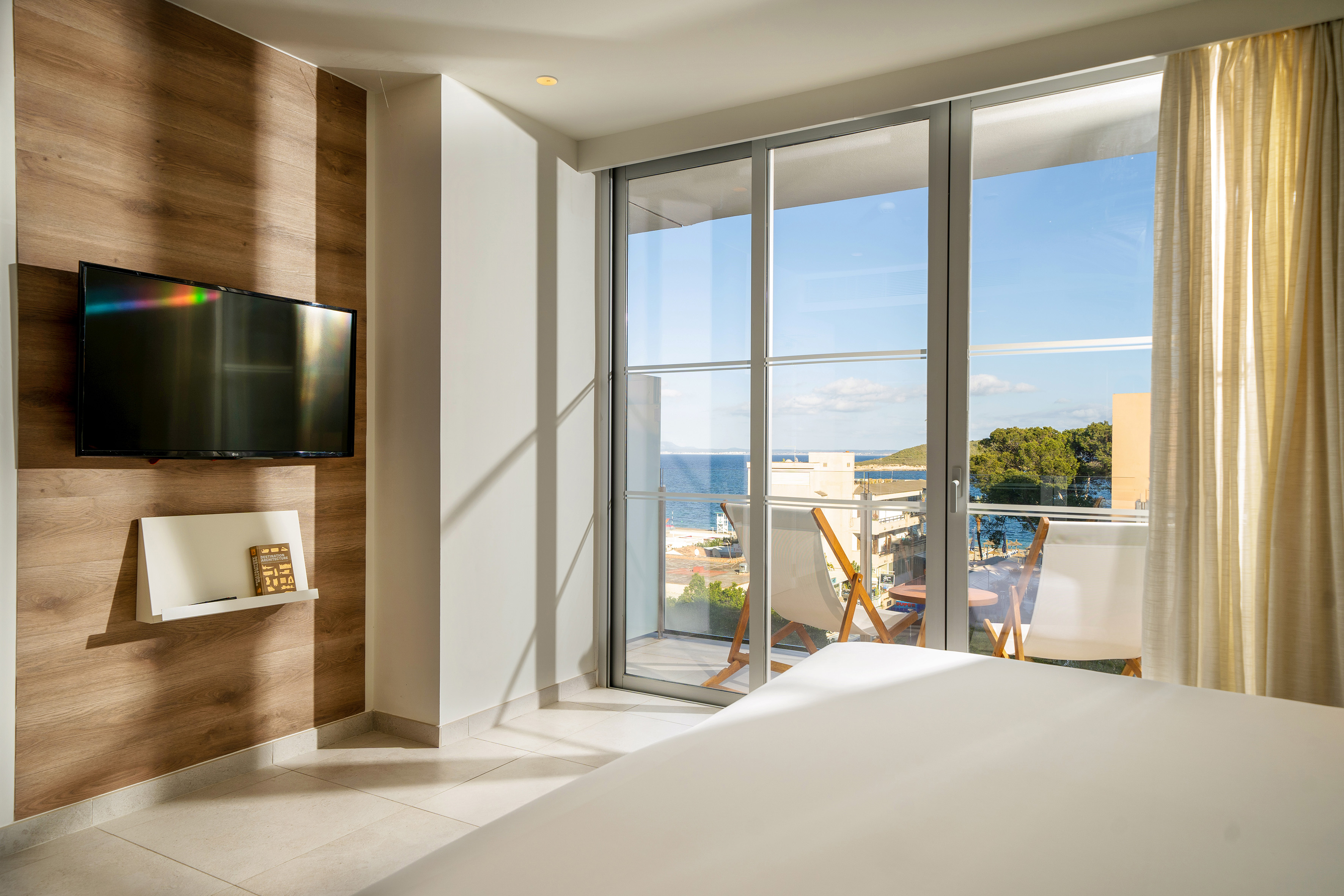 Bright modern hotel room with sea view balcony, large bed, and wall-mounted TV.