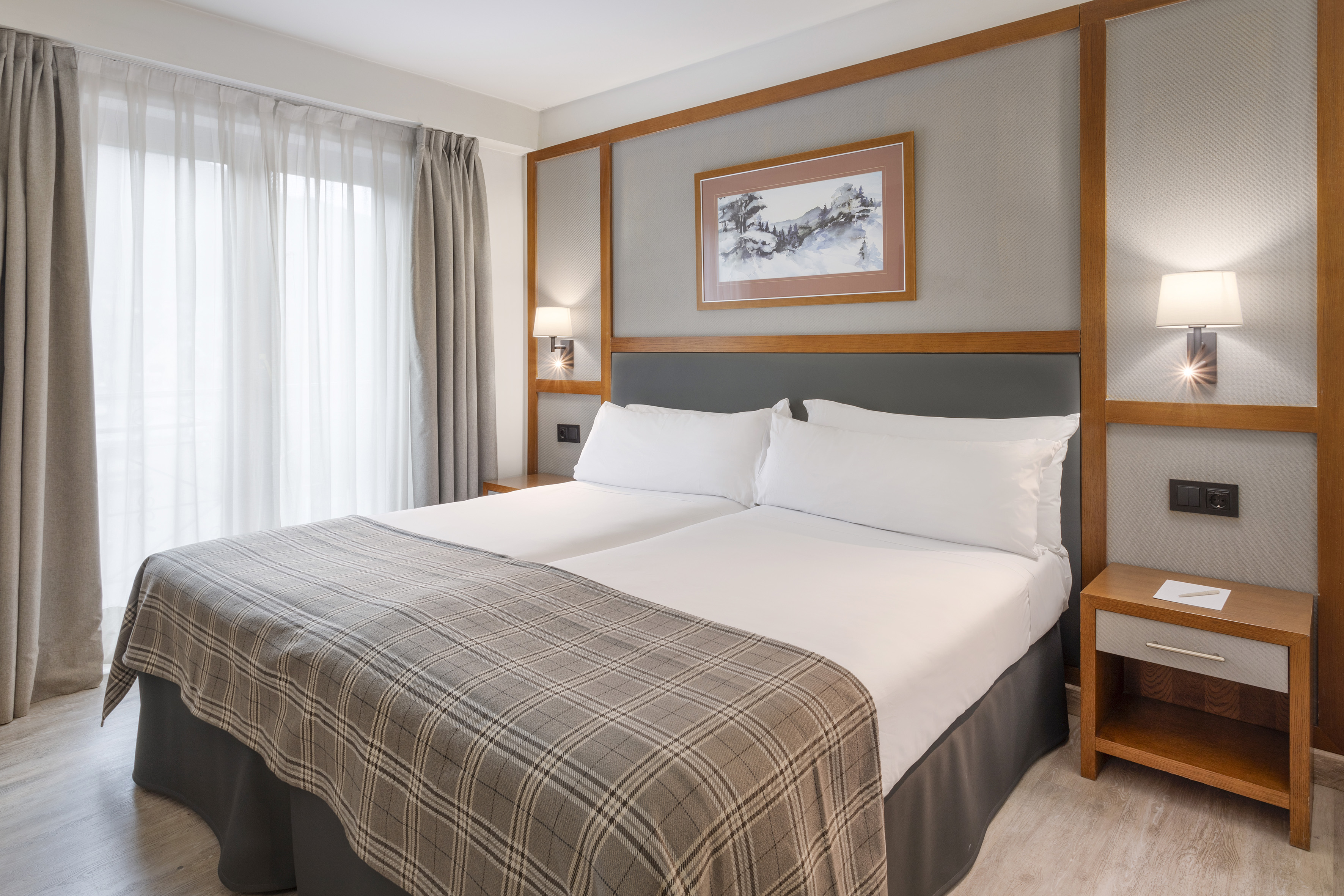 Hotel guest room with two beds, plaid blanket, and sheer window curtains.