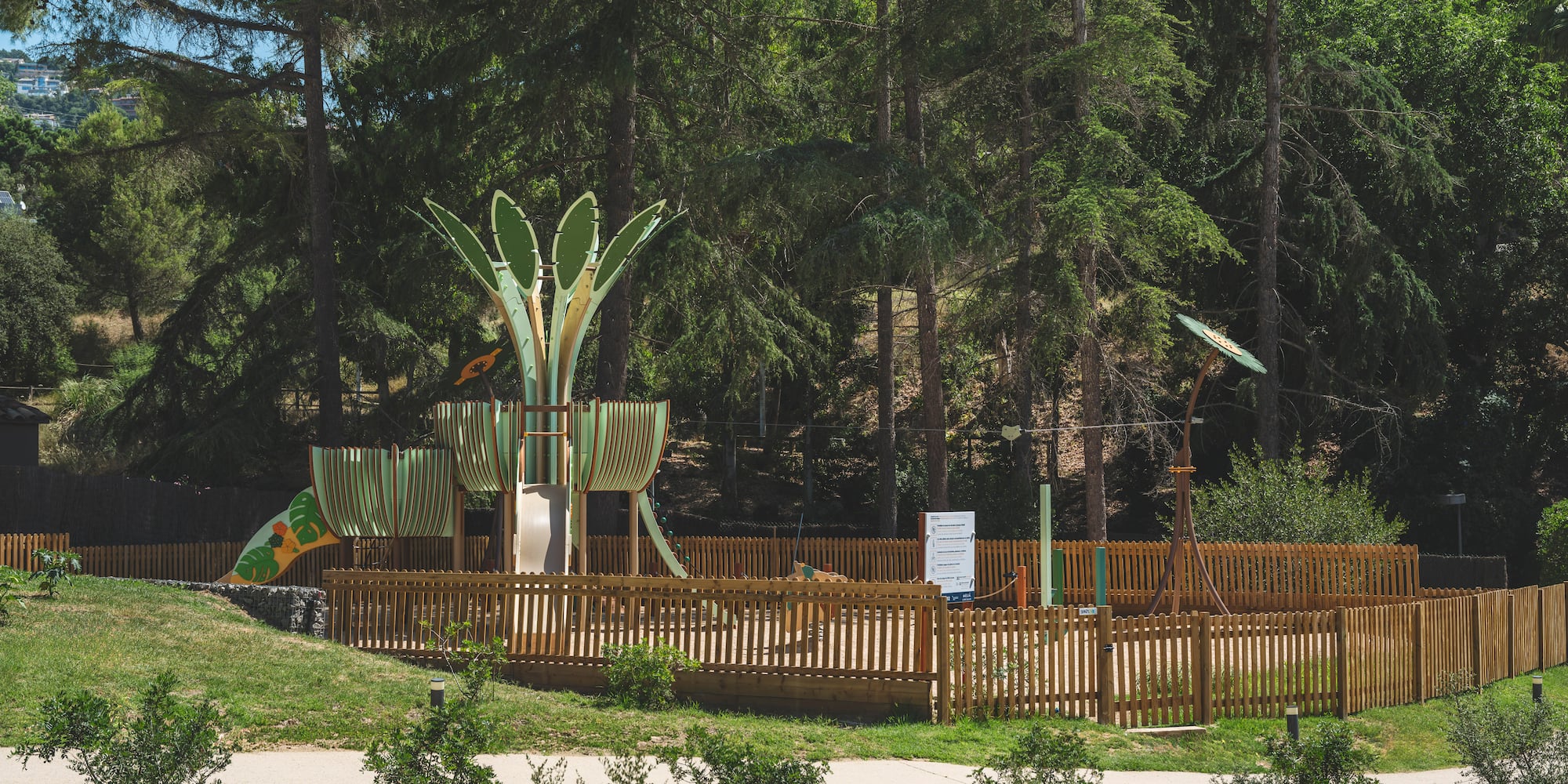 Fenced outdoor playground with green slide and climbing structures among pine trees.