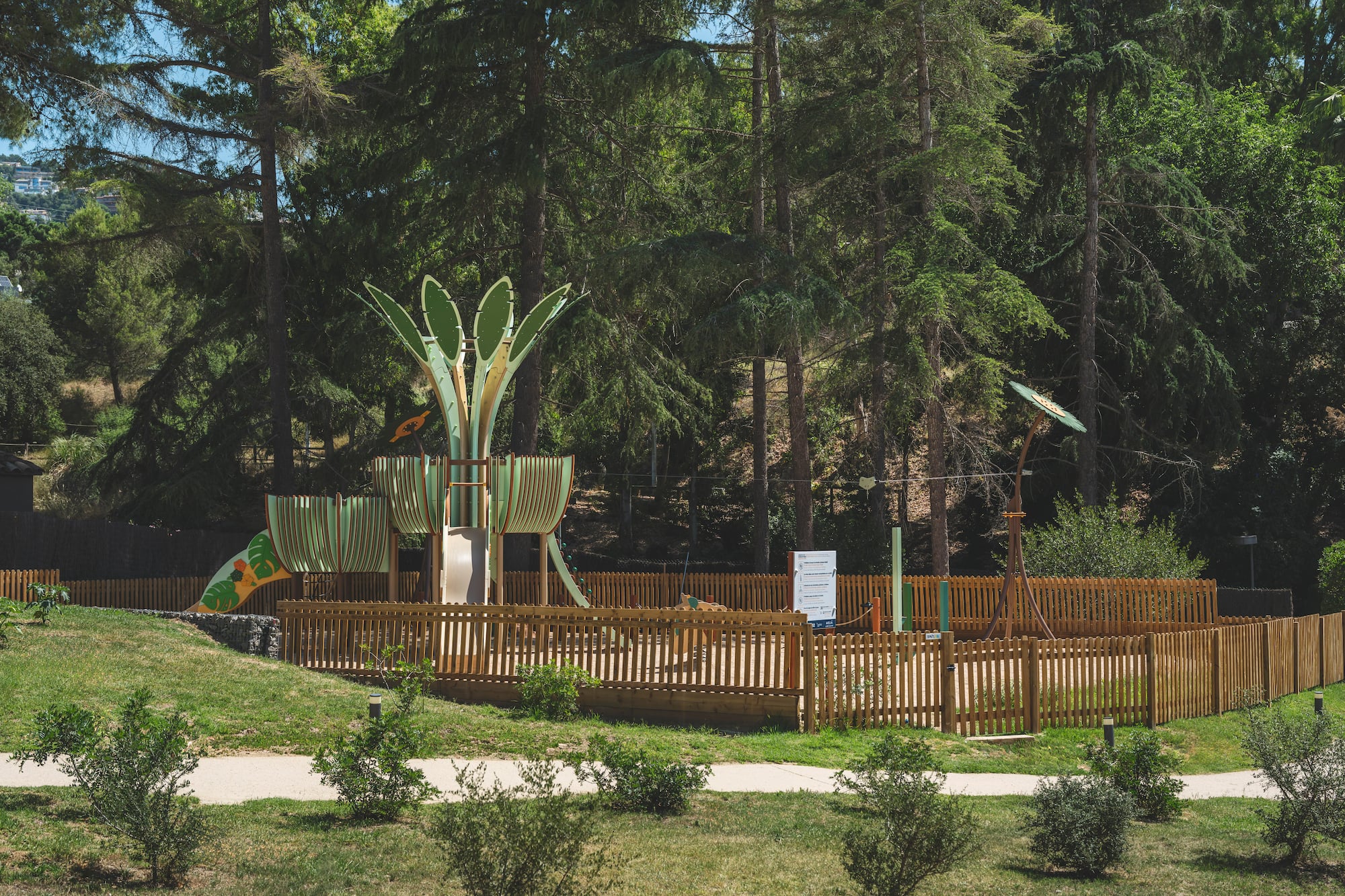 Fenced outdoor playground with green slide and climbing structures among pine trees.