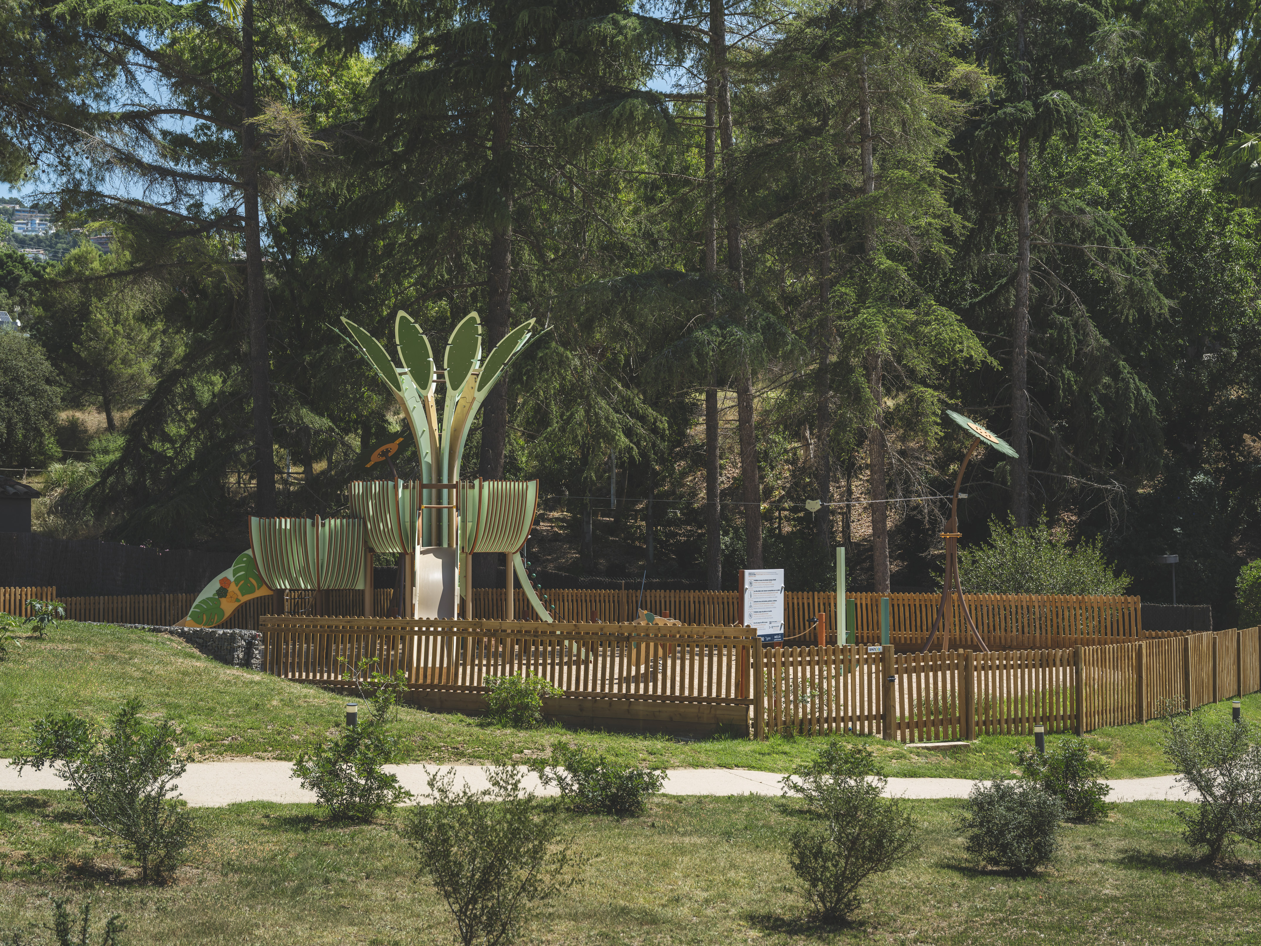 Fenced outdoor playground with green slide and climbing structures among pine trees.