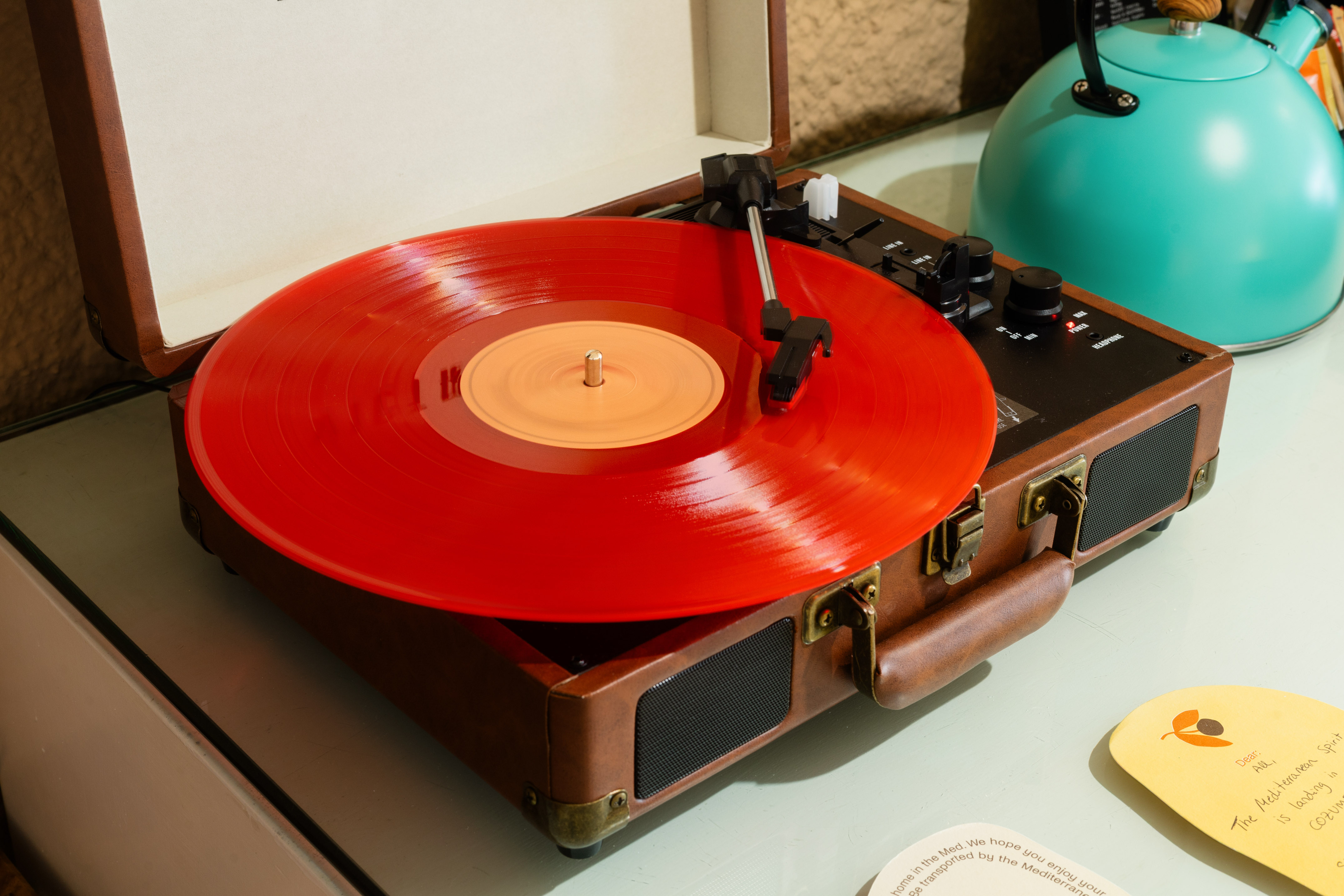 Tocadiscos de maleta con vinilo rojo. Detrás, tetera turquesa.