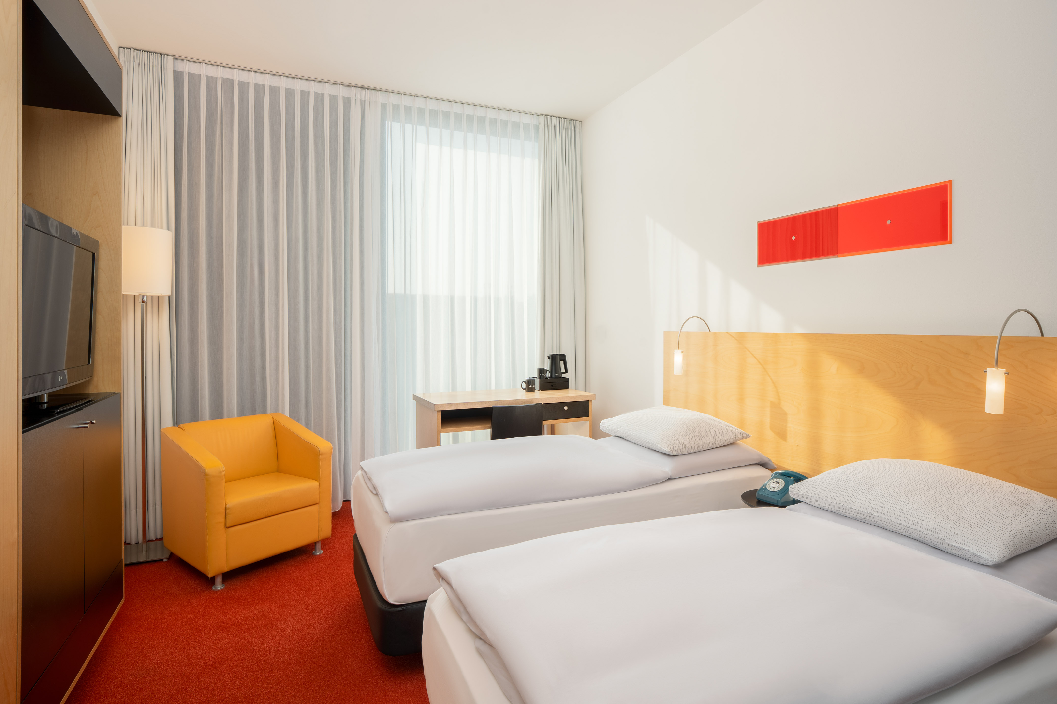 Hotel room with two single beds, orange armchair, desk, and red carpet.