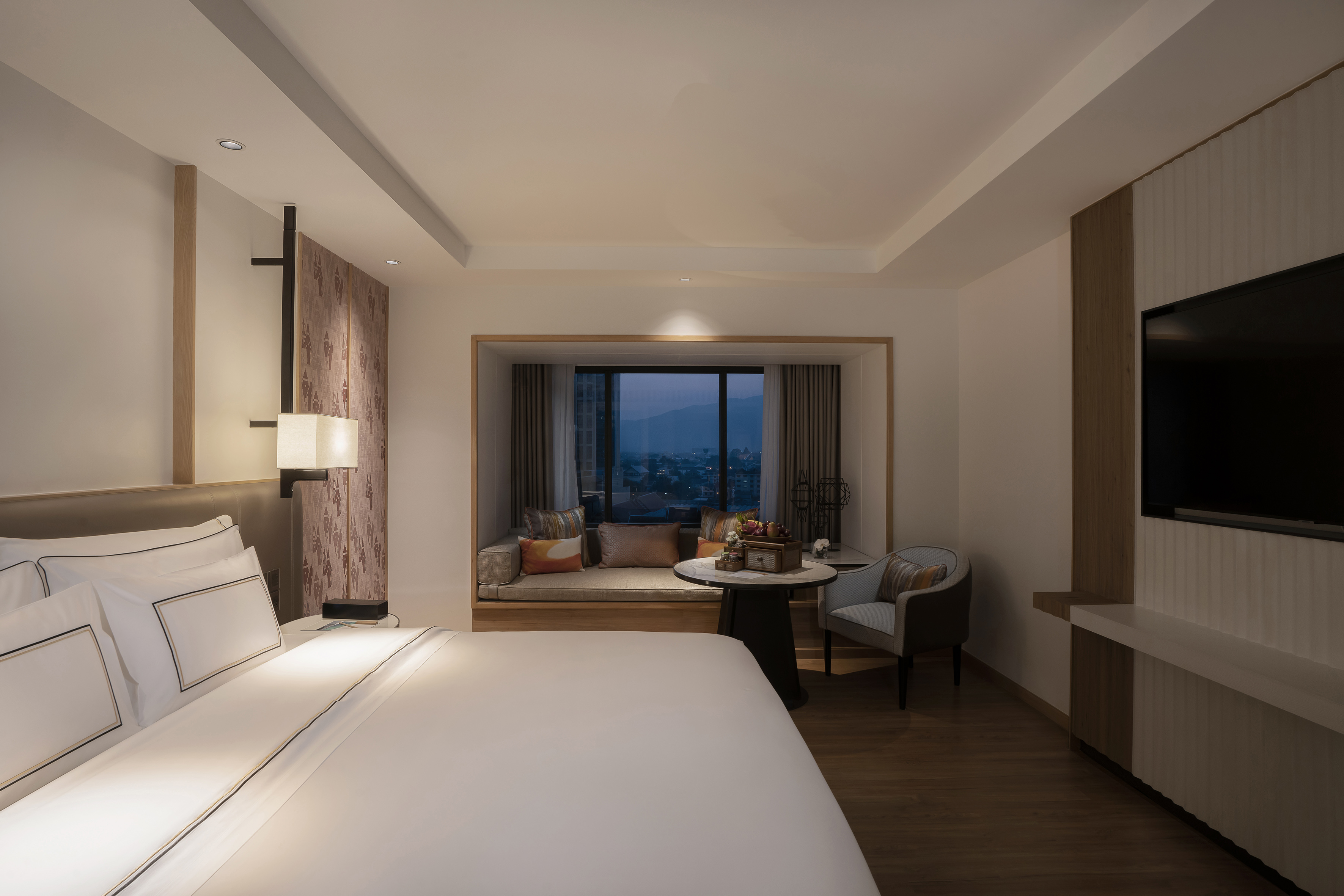 Modern hotel room with large bed, bay window, evening mountain view.