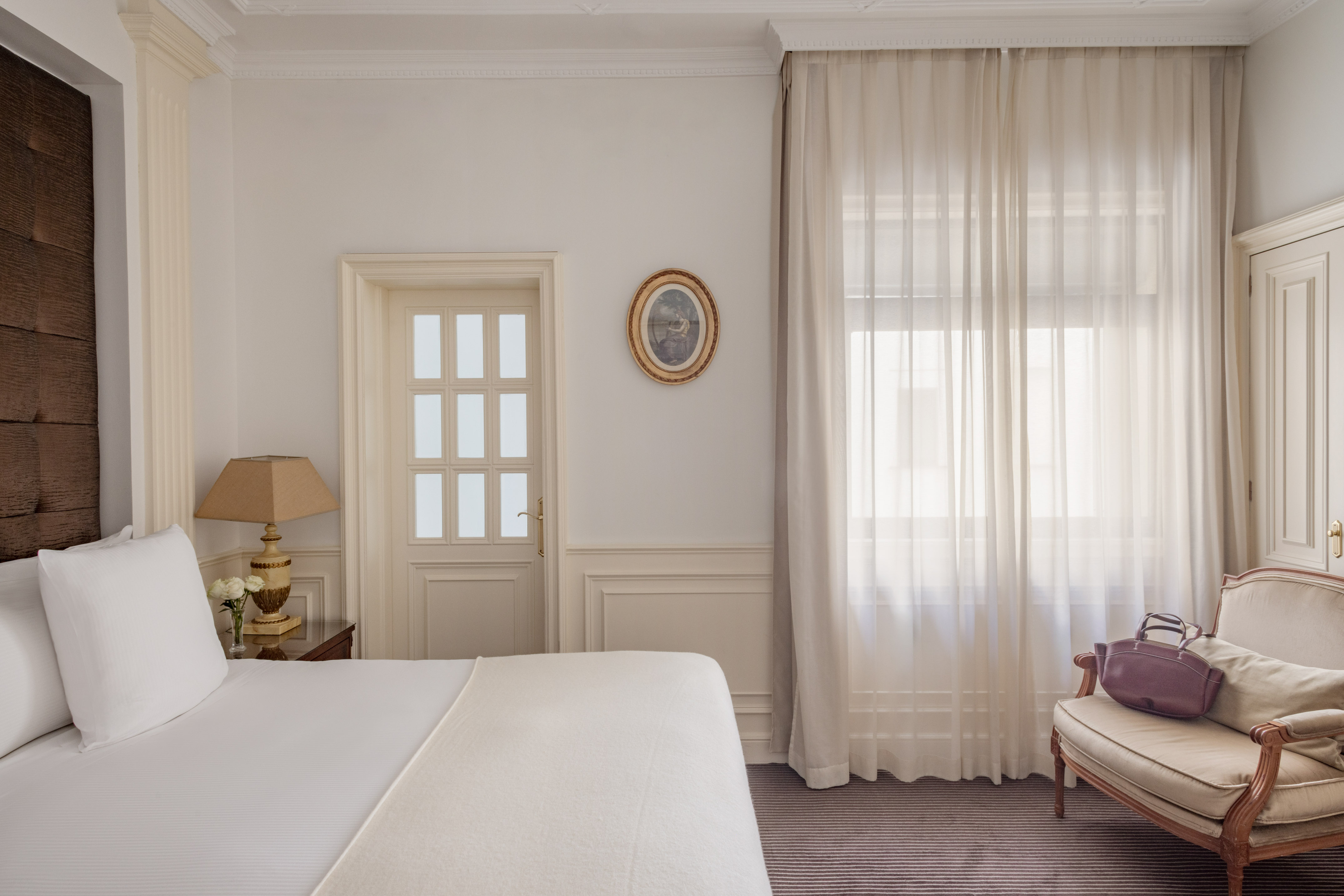 Elegant hotel bedroom with white bed, classic armchair, and sheer curtains.