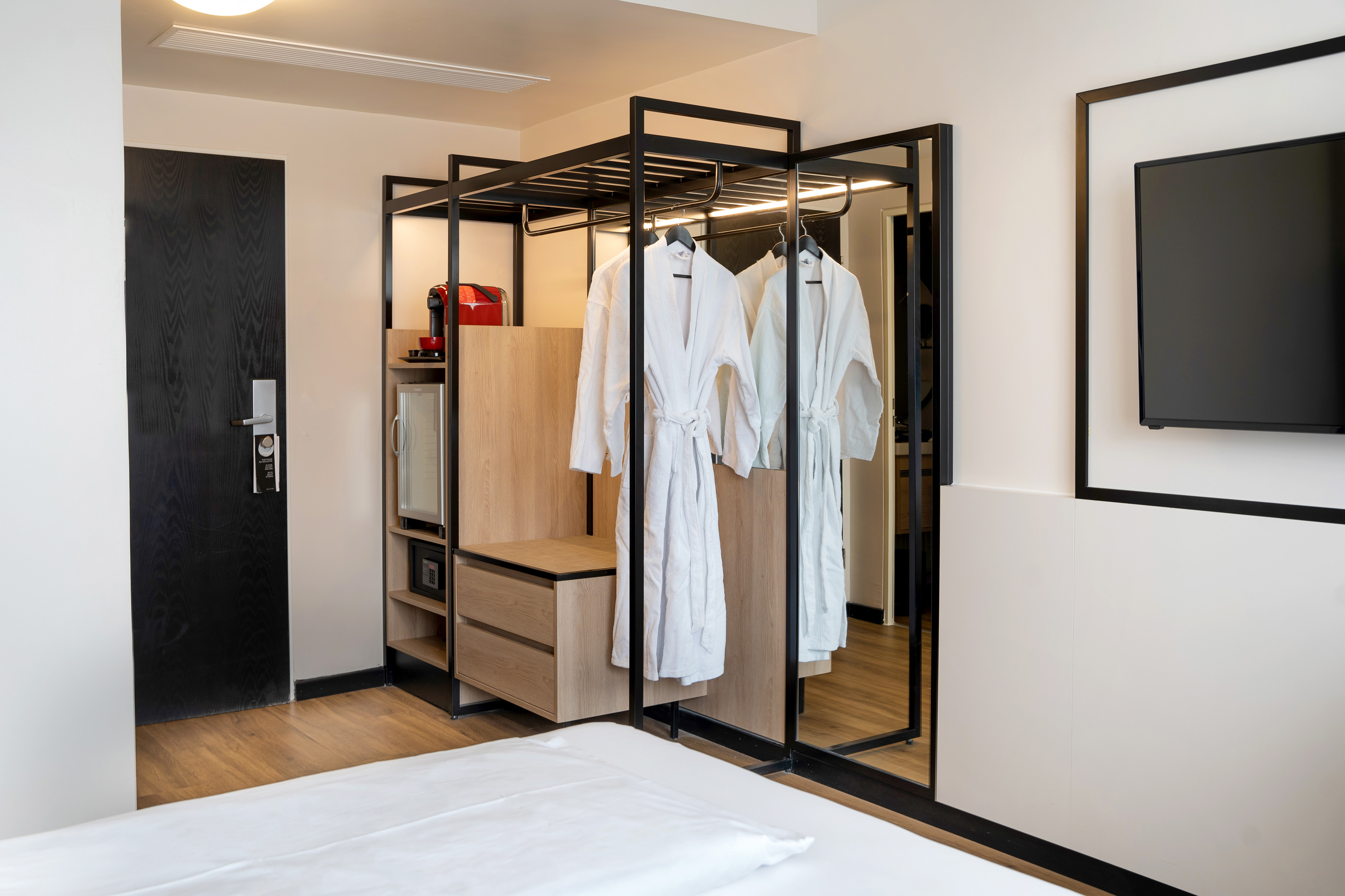 Modern hotel room with open wardrobe, bathrobes, Nespresso, mini-fridge, and TV.