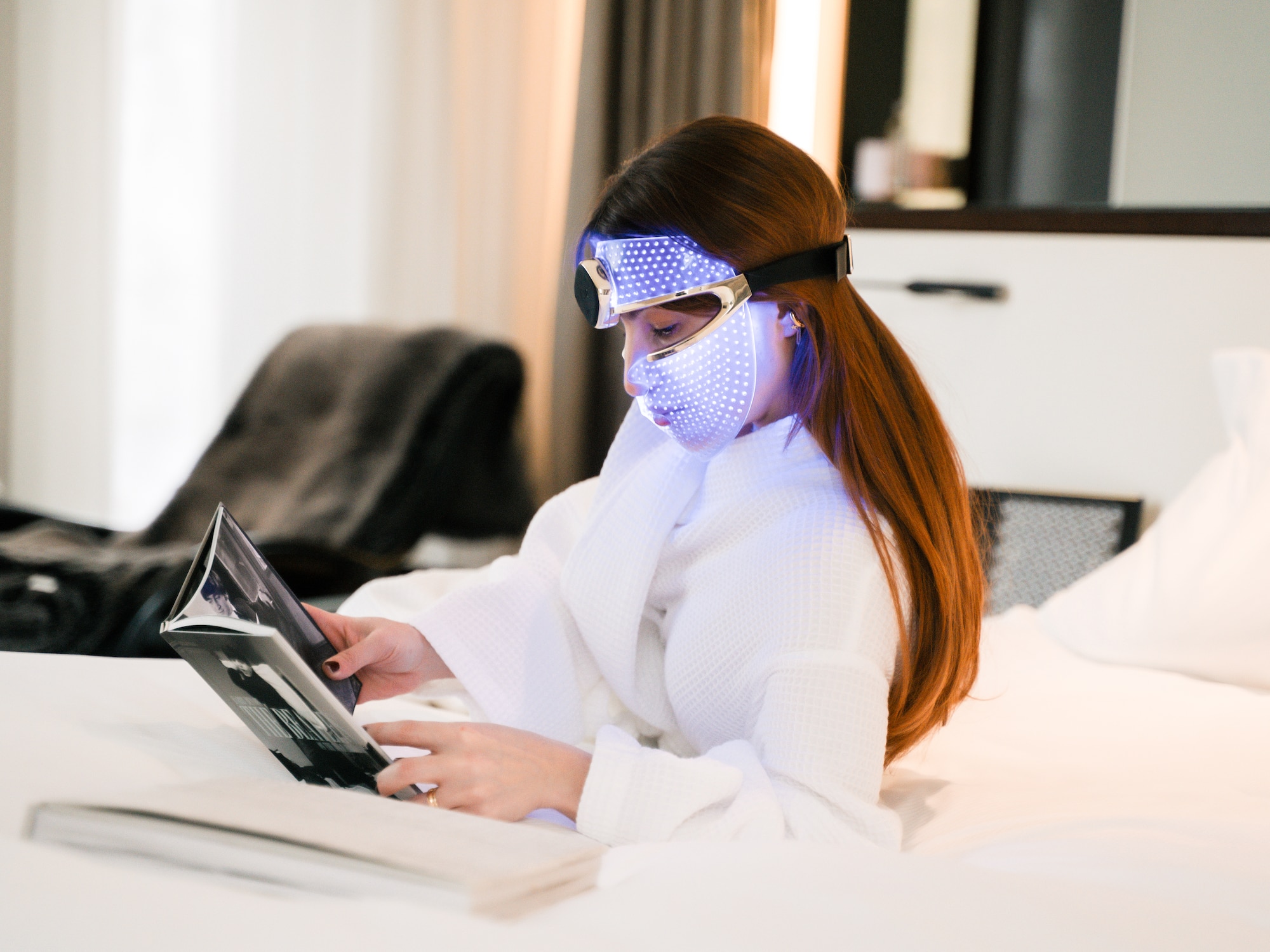 Woman in LED therapy mask reads. Bathrobe, skincare, beauty tech relaxation.