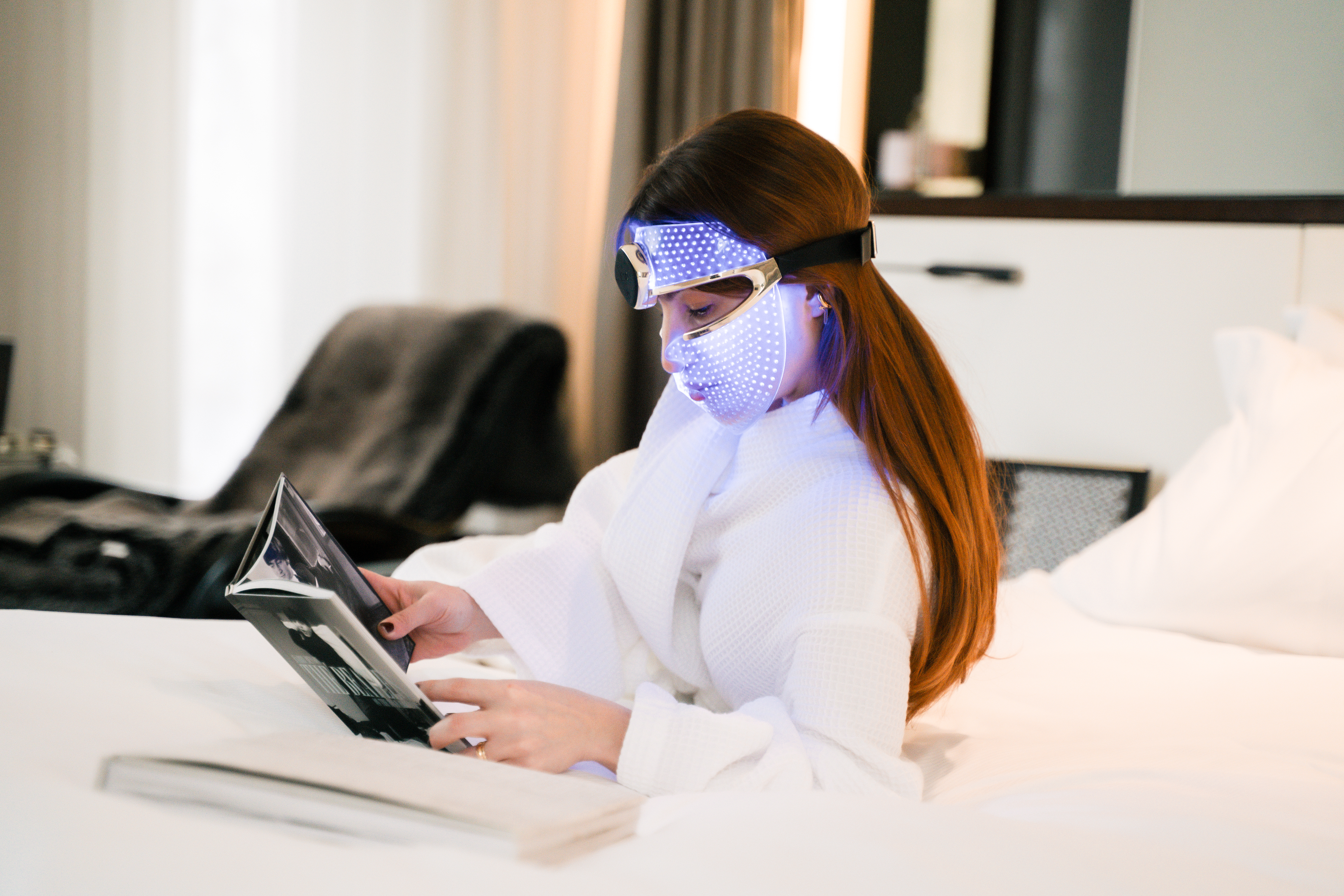 Woman in LED therapy mask reads. Bathrobe, skincare, beauty tech relaxation.