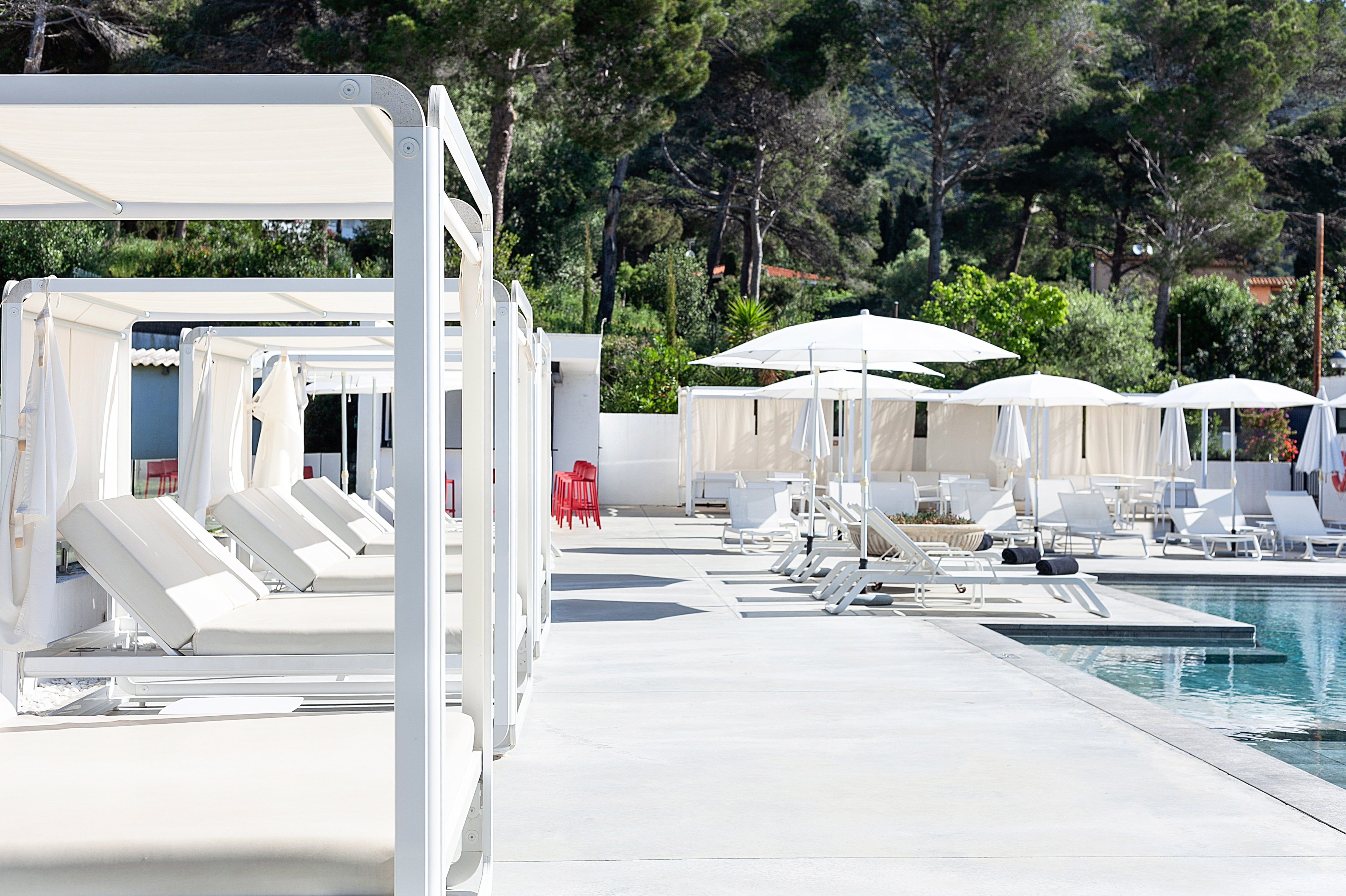 Modern resort pool with white cabanas, loungers, umbrellas, and greenery.