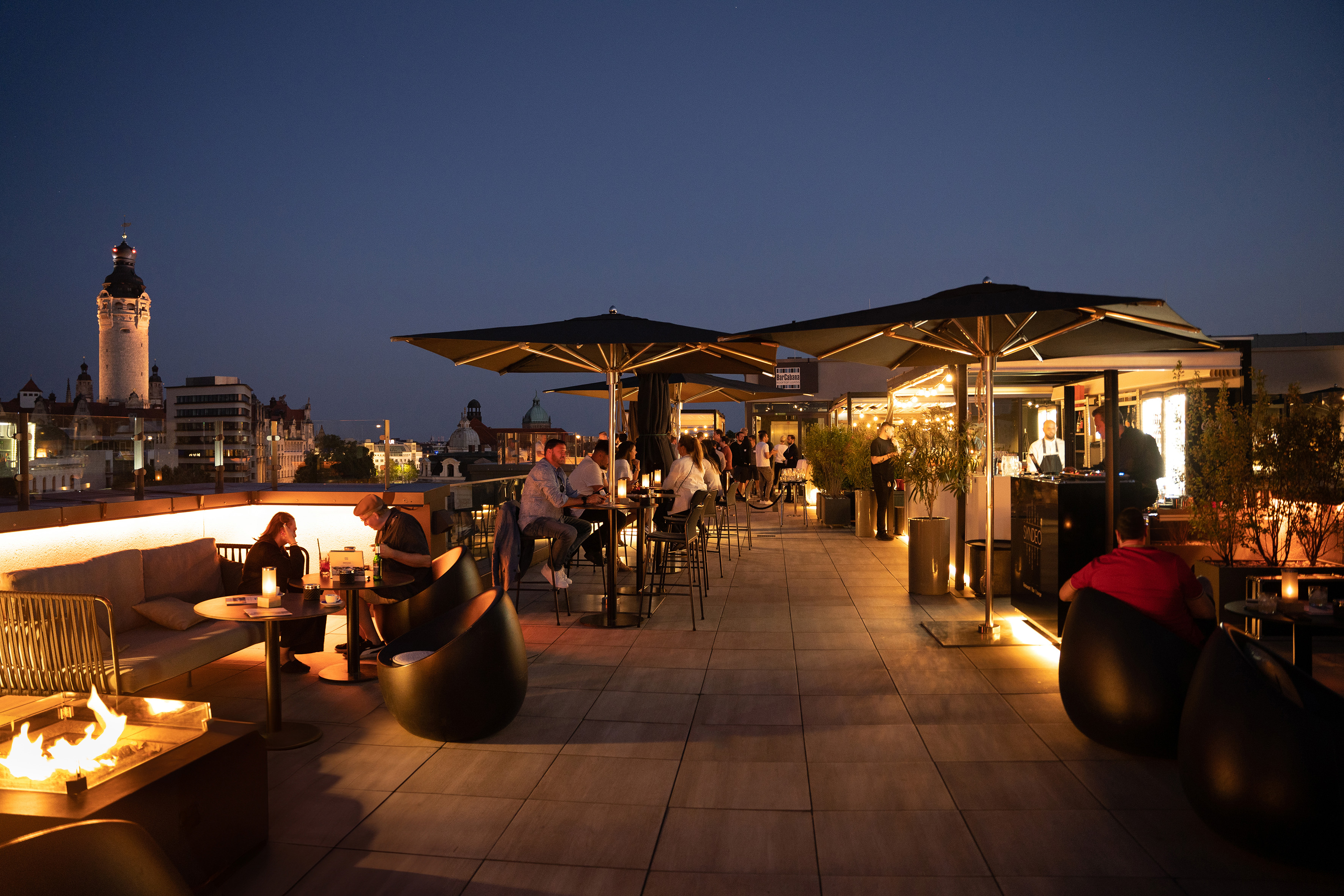 Leipzig rooftop bar at dusk with patrons, illuminated city skyline, fire pit.