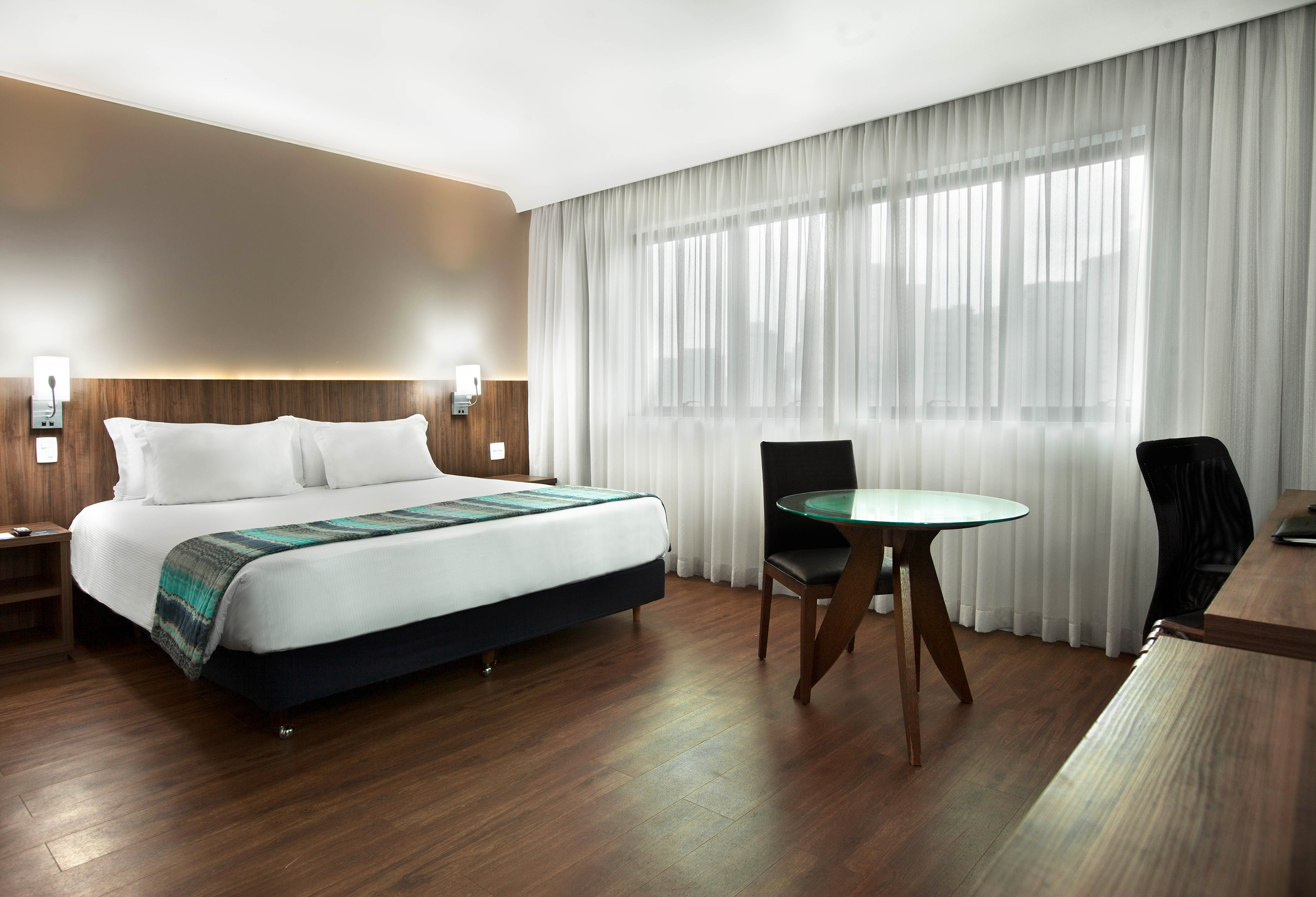 Hotel room with king bed, wood floor, glass table, chairs, and large window.