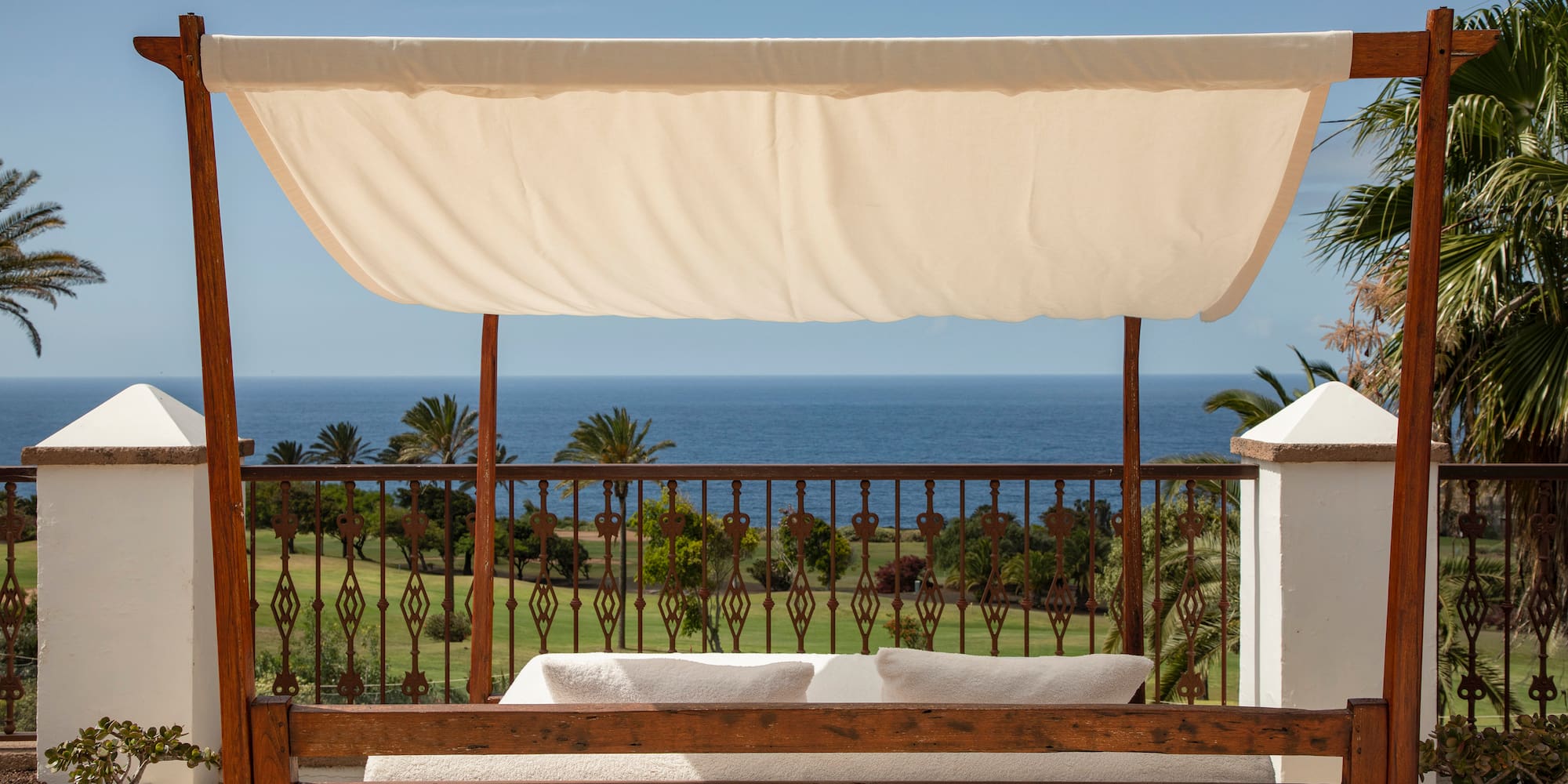 Wooden daybed with canopy overlooking ocean and green golf course from a terrace.