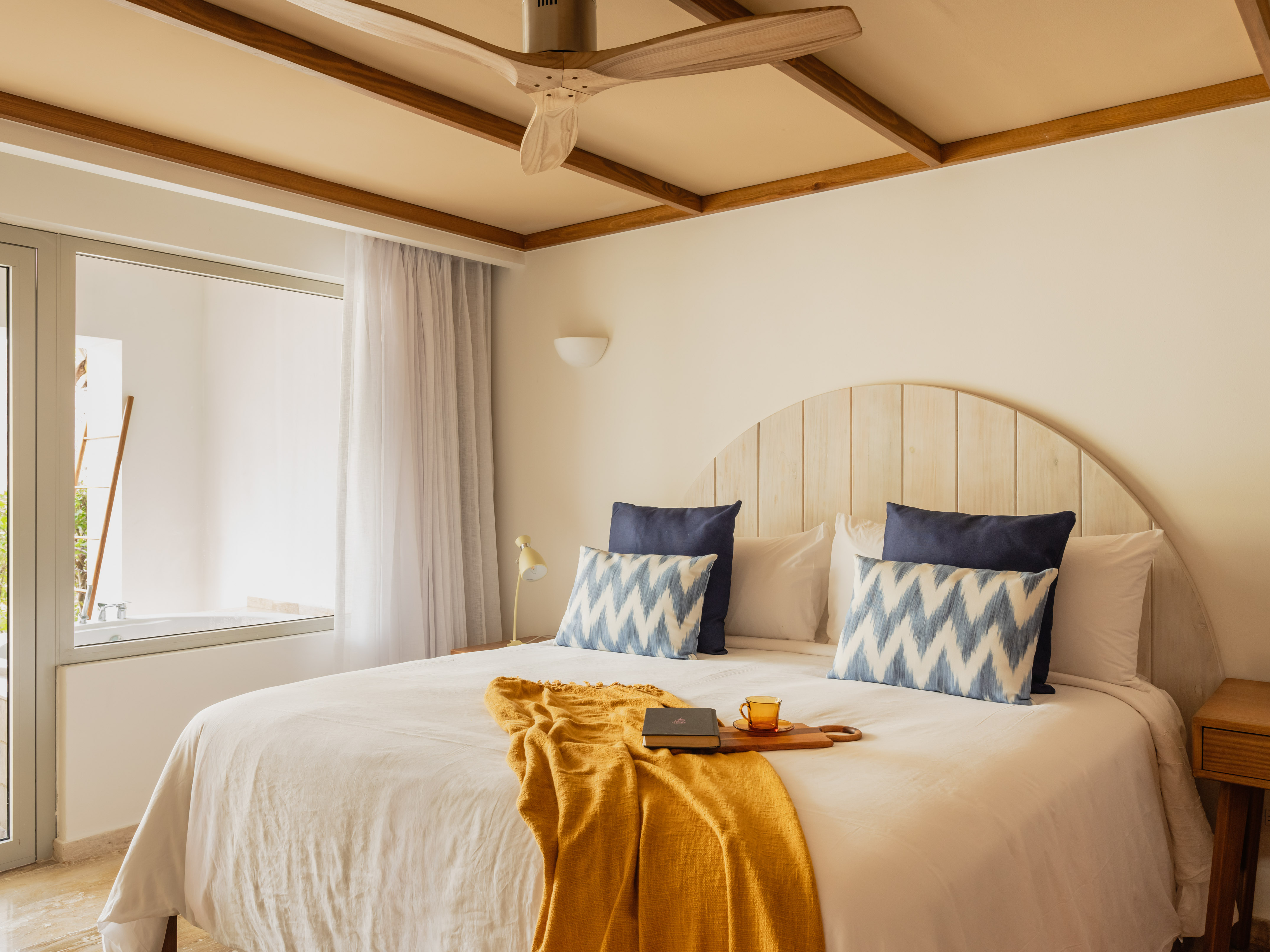 Modern resort bedroom with white bed, blue pillows, yellow throw, wooden fan.