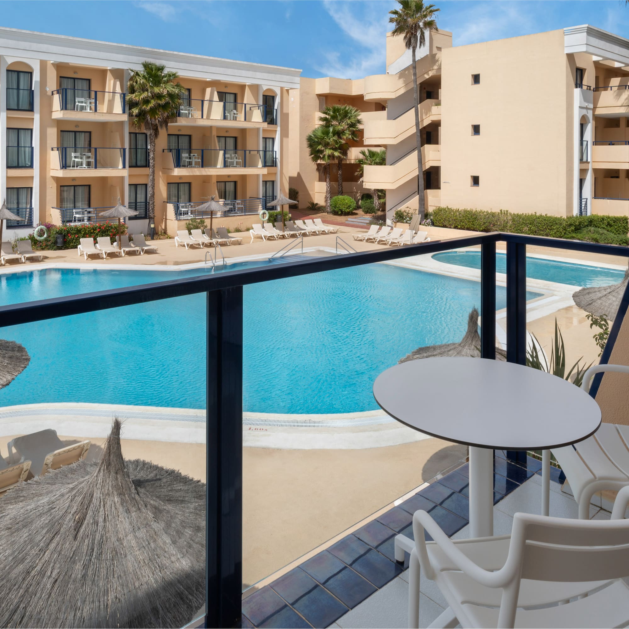 Sunny hotel resort with large swimming pool, loungers, and palm trees.