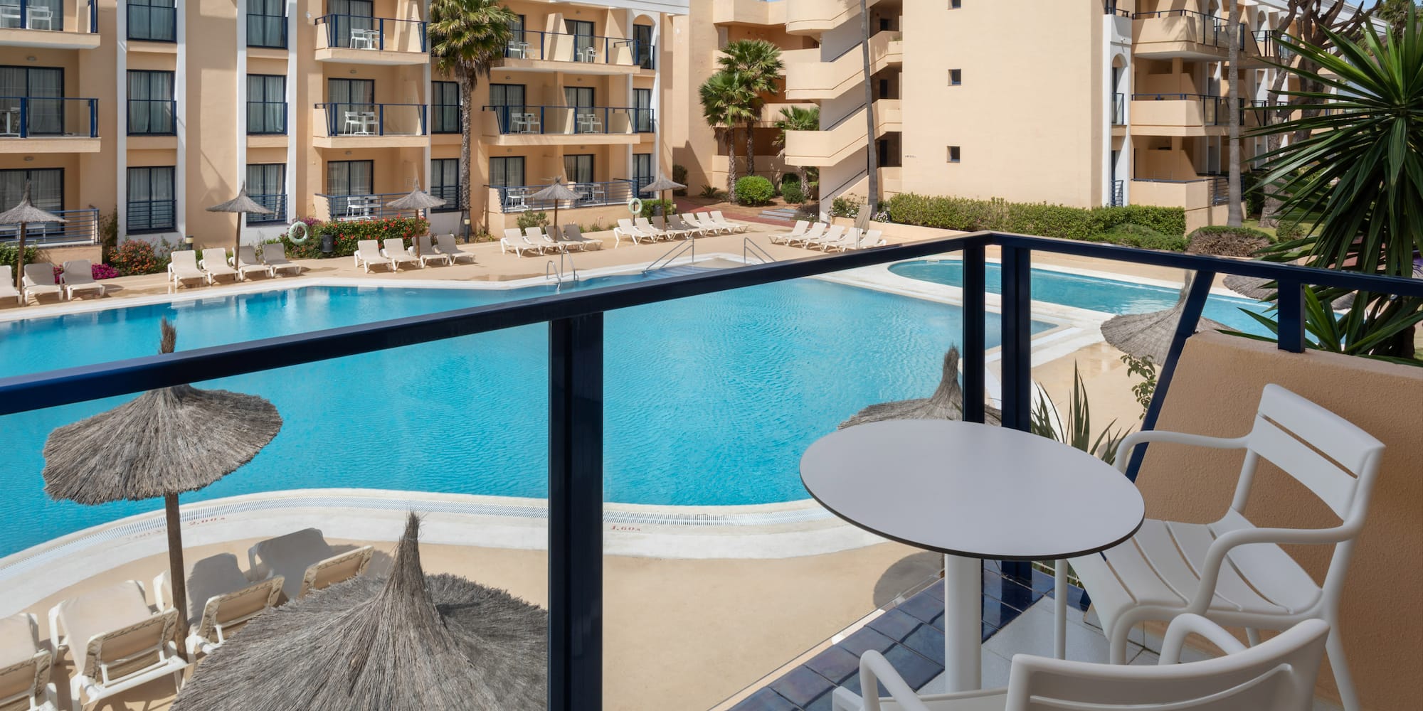 Sunny hotel resort with large swimming pool, loungers, and palm trees.