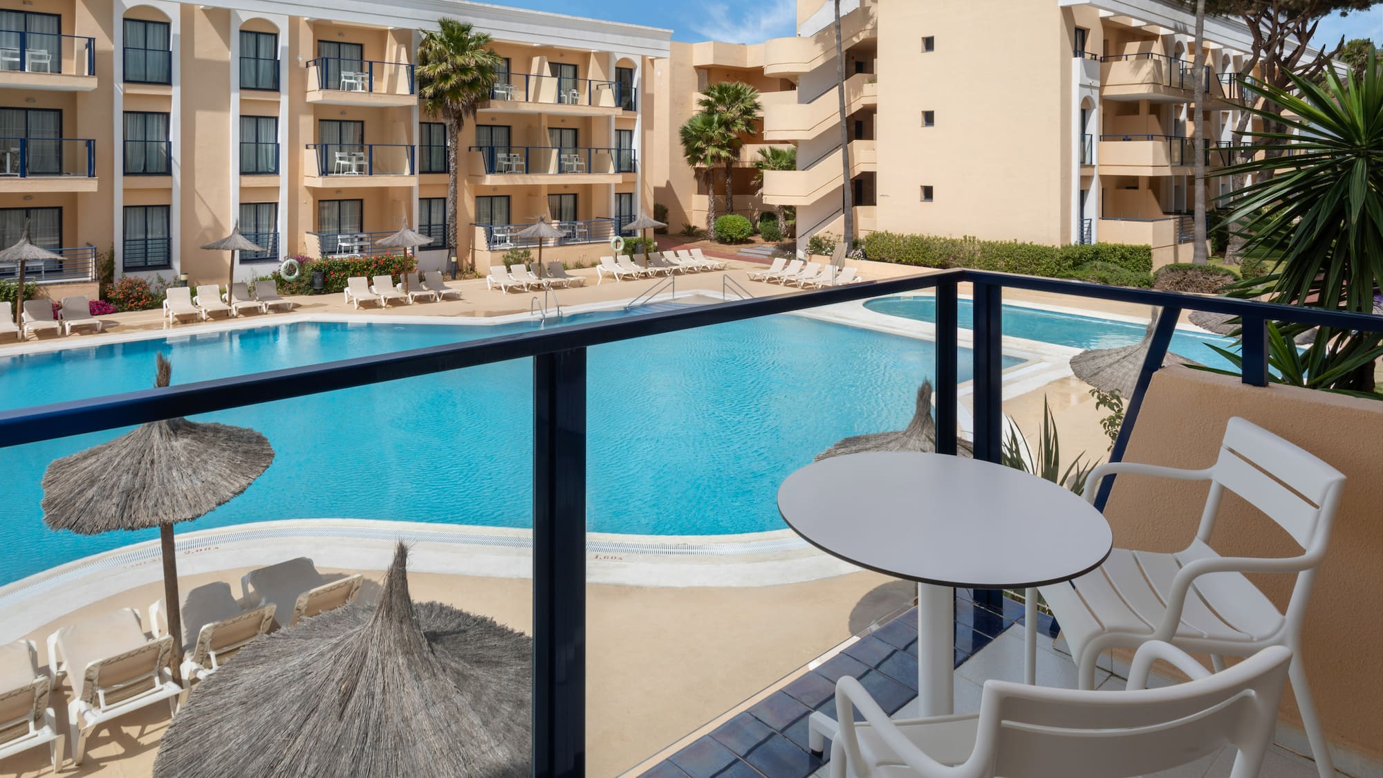 Balcony view of resort hotel with large swimming pool, sun loungers, and palm trees.