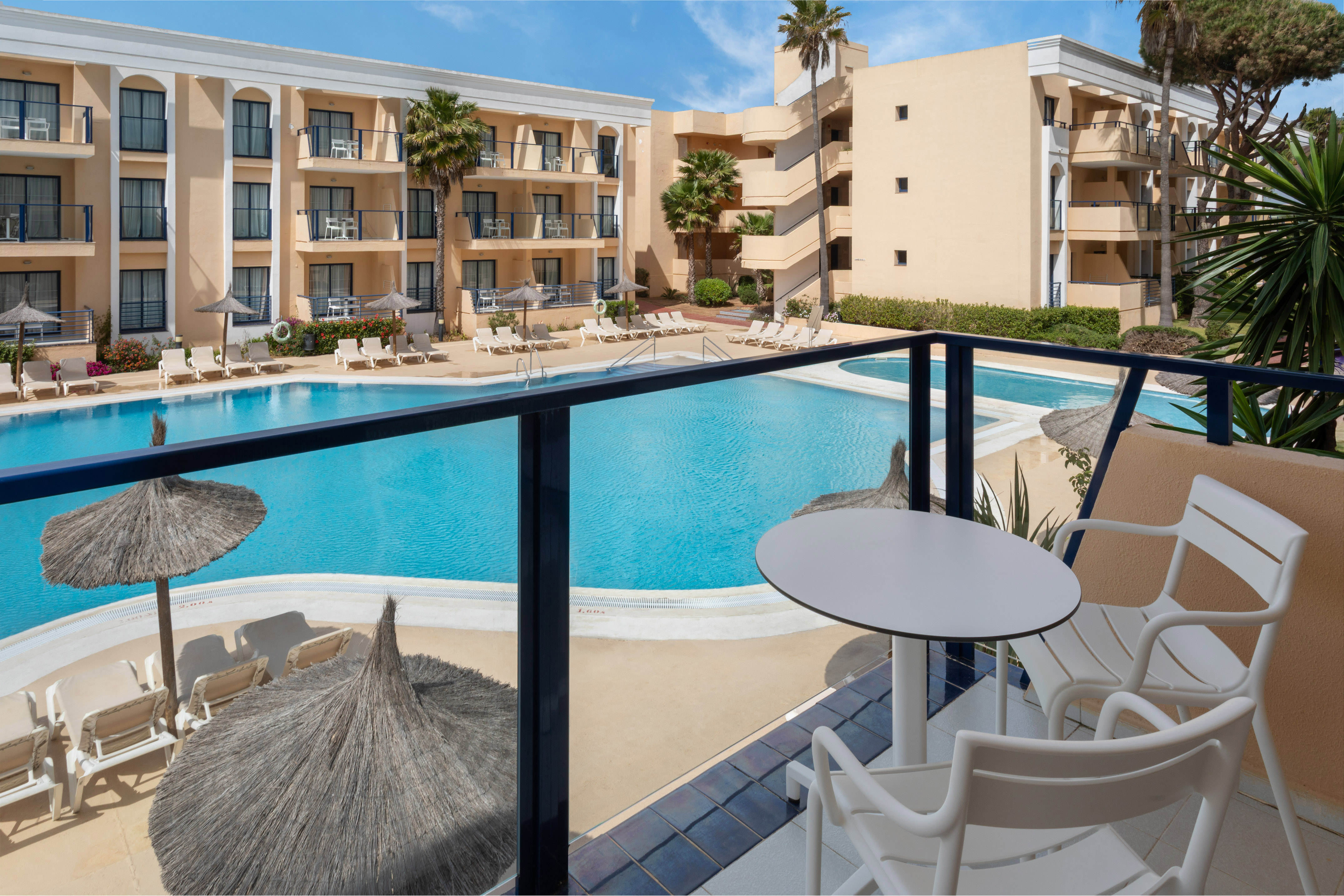 Balcony view of resort hotel with large swimming pool, sun loungers, and palm trees.