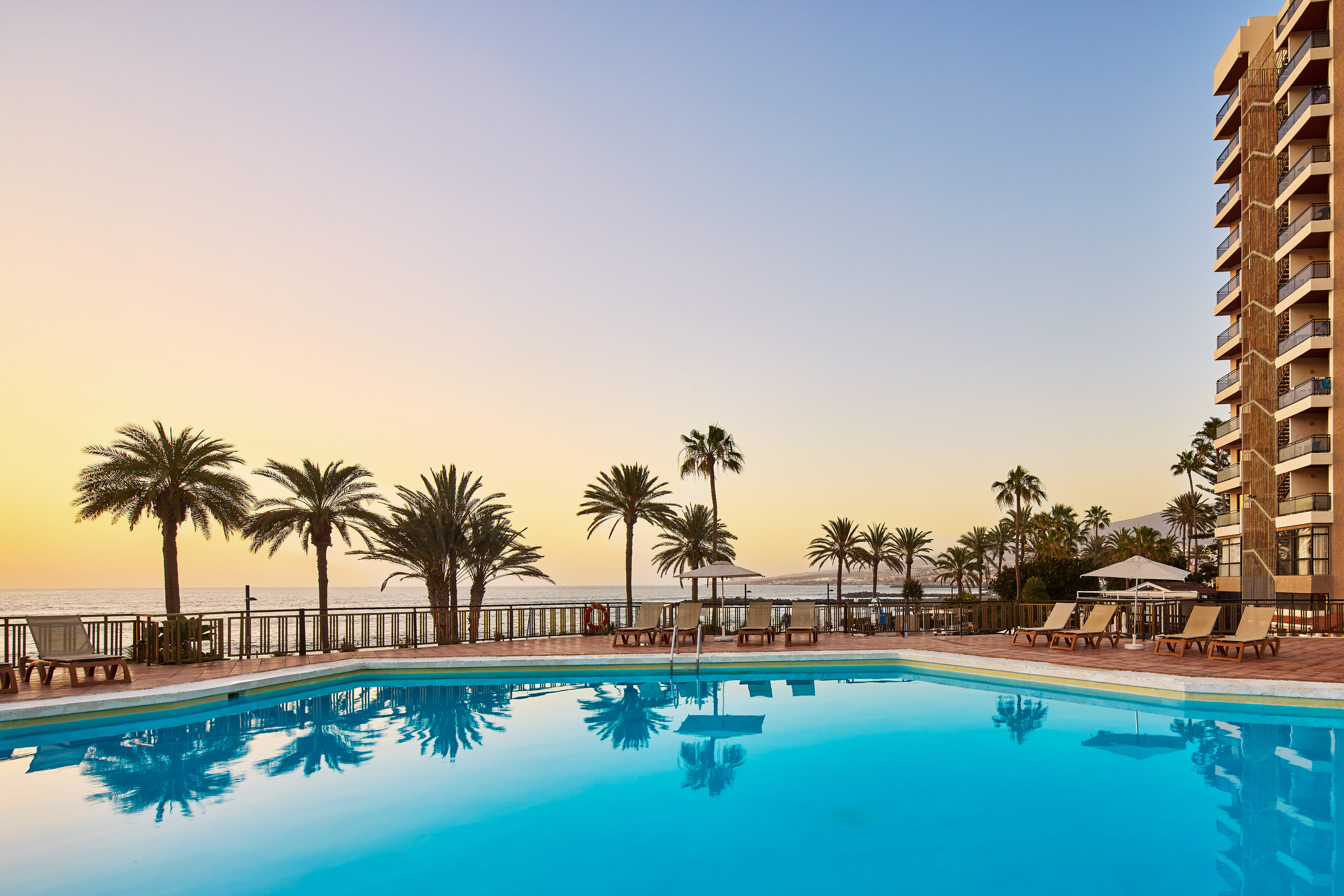 Tropical resort pool at sunset with palm trees, ocean view, and hotel building.