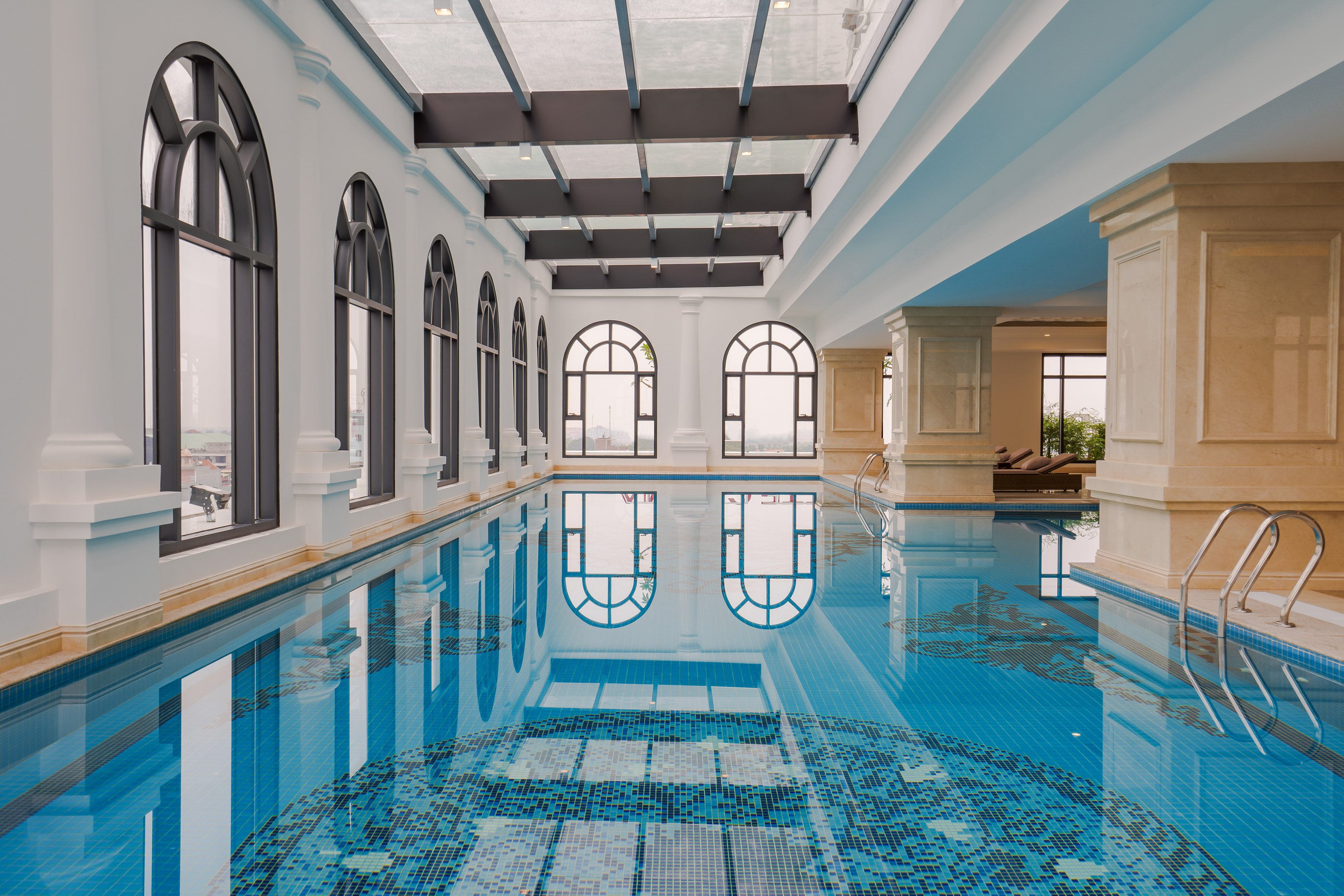 Luxury indoor hotel pool with arched windows, skylight, and blue mosaic.