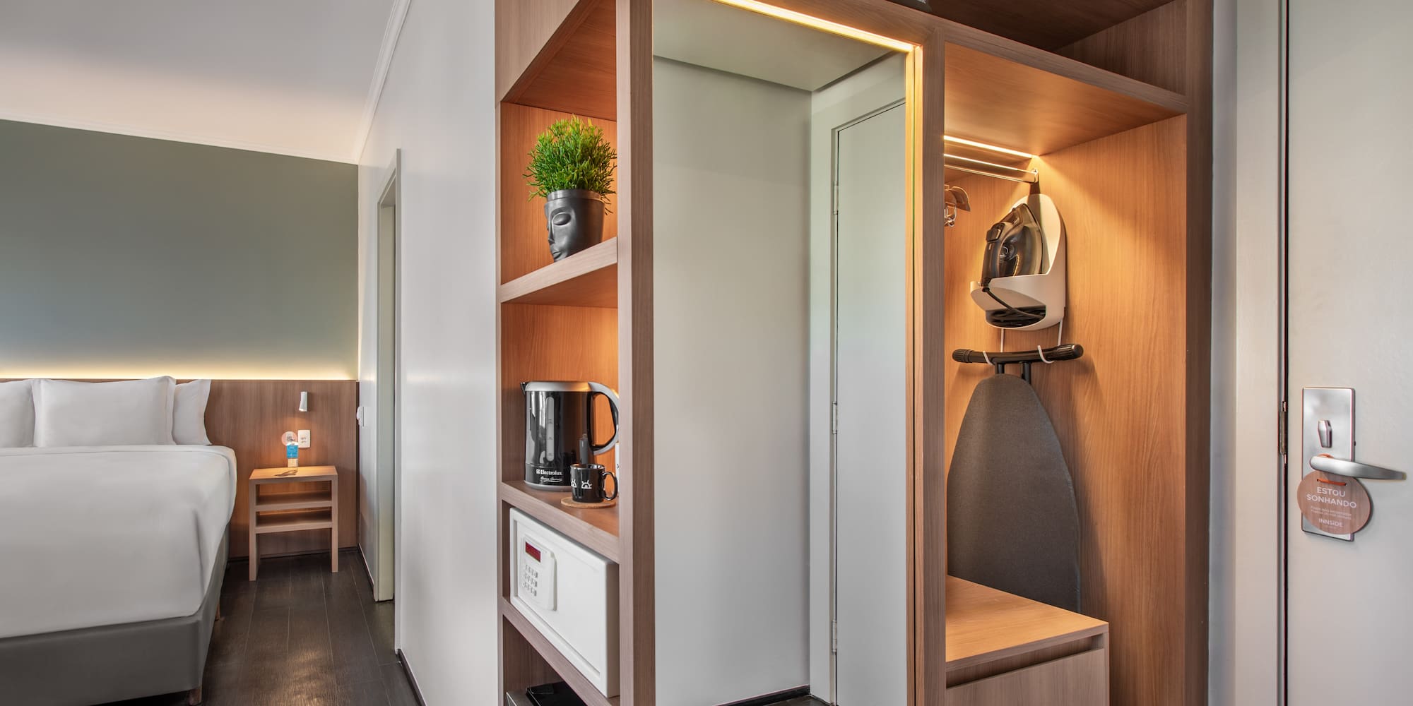 Modern hotel room with bed, wooden closet, ironing board, safe, and kettle.