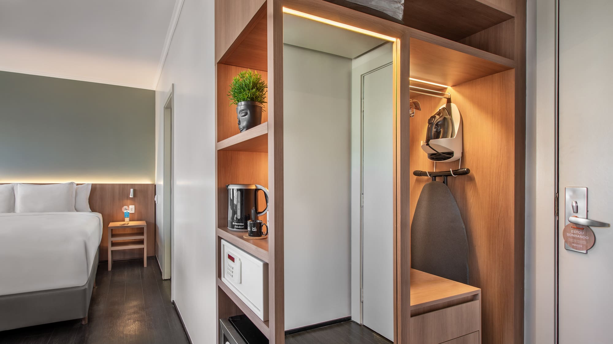Modern hotel room with bed, wooden closet, ironing board, safe, and kettle.