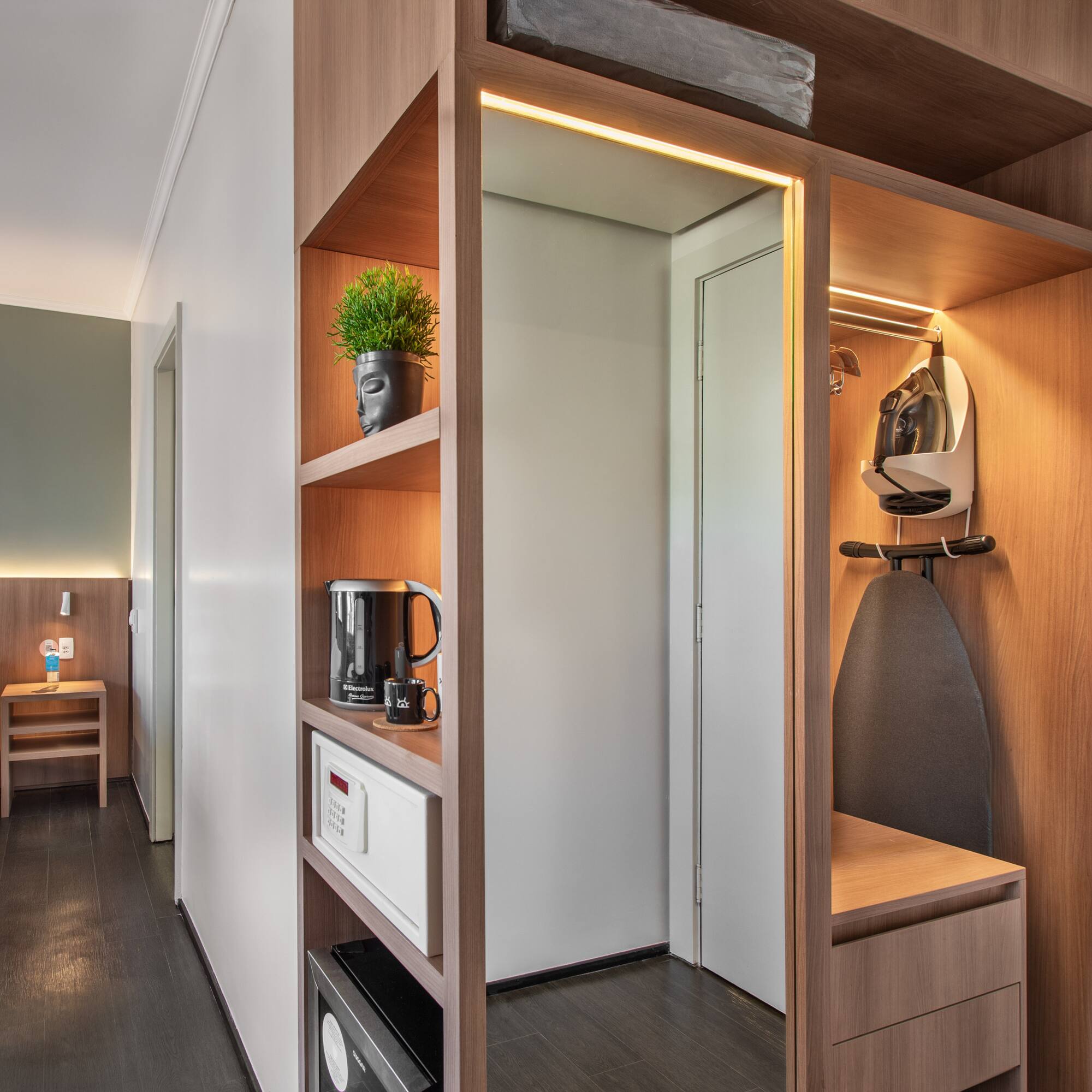 Modern hotel room with bed, wooden closet, ironing board, safe, and kettle.
