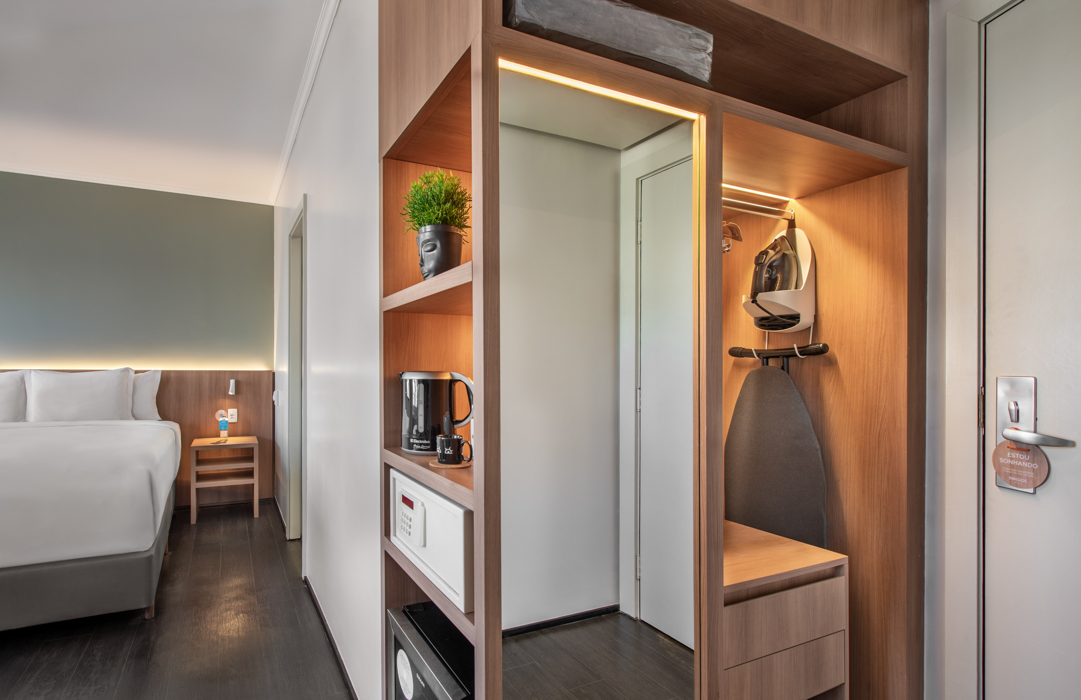 Modern hotel room with bed, wooden closet, ironing board, safe, and kettle.
