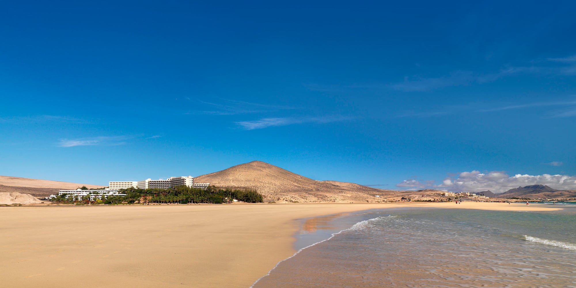 Extensa playa de arena, mar, resort y montañas áridas bajo cielo azul.