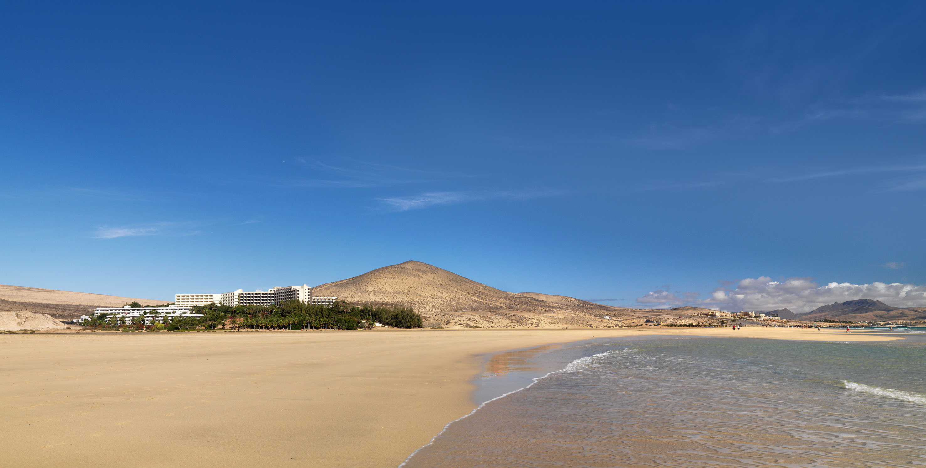 Extensa playa de arena, mar, resort y montañas áridas bajo cielo azul.