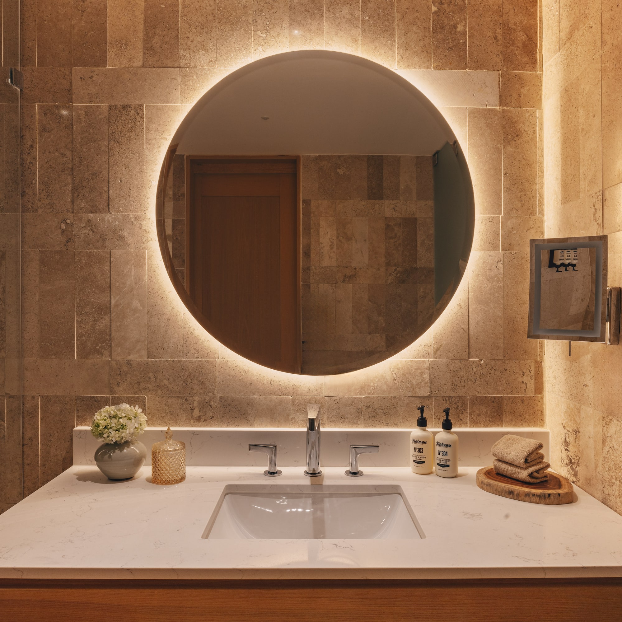 Modern bathroom vanity with backlit round mirror, stone wall, and white sink.