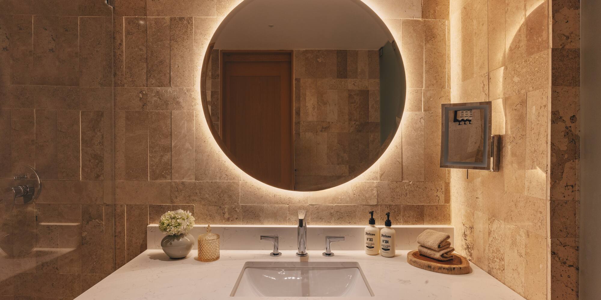 Modern bathroom vanity with backlit round mirror, stone wall, and white sink.