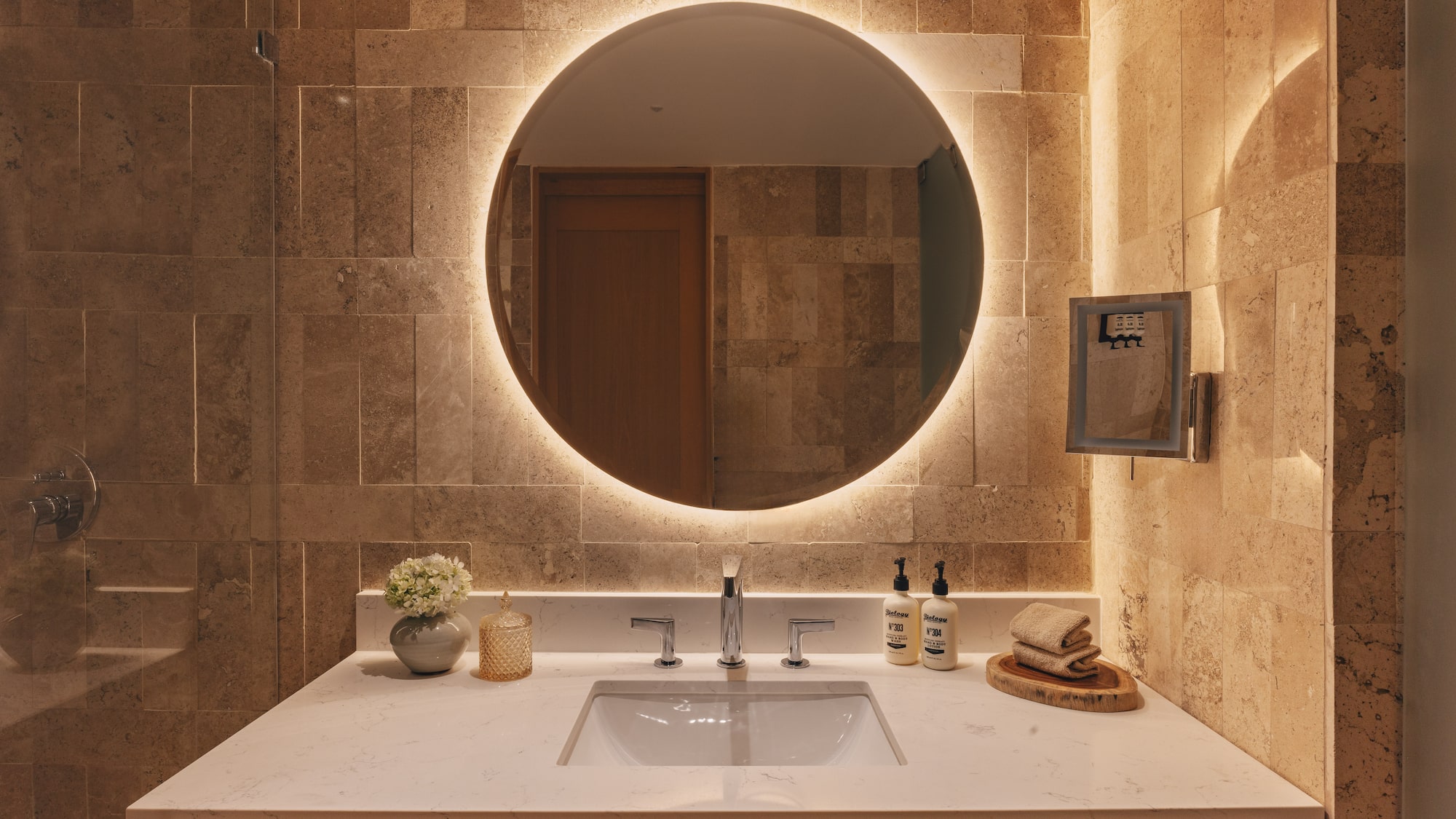 Modern bathroom vanity with backlit round mirror, stone wall, and white sink.
