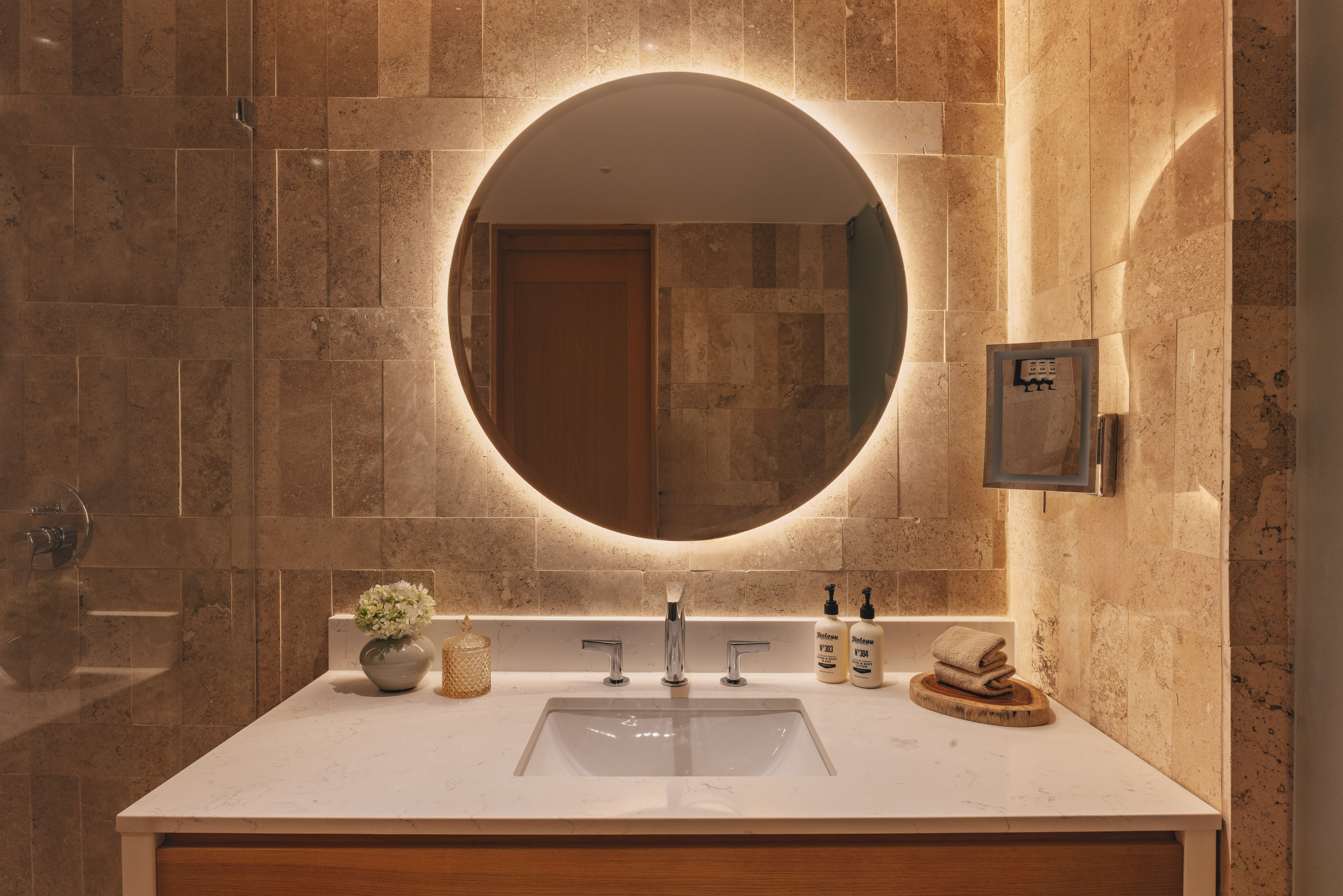 Modern bathroom vanity with backlit round mirror, stone wall, and white sink.