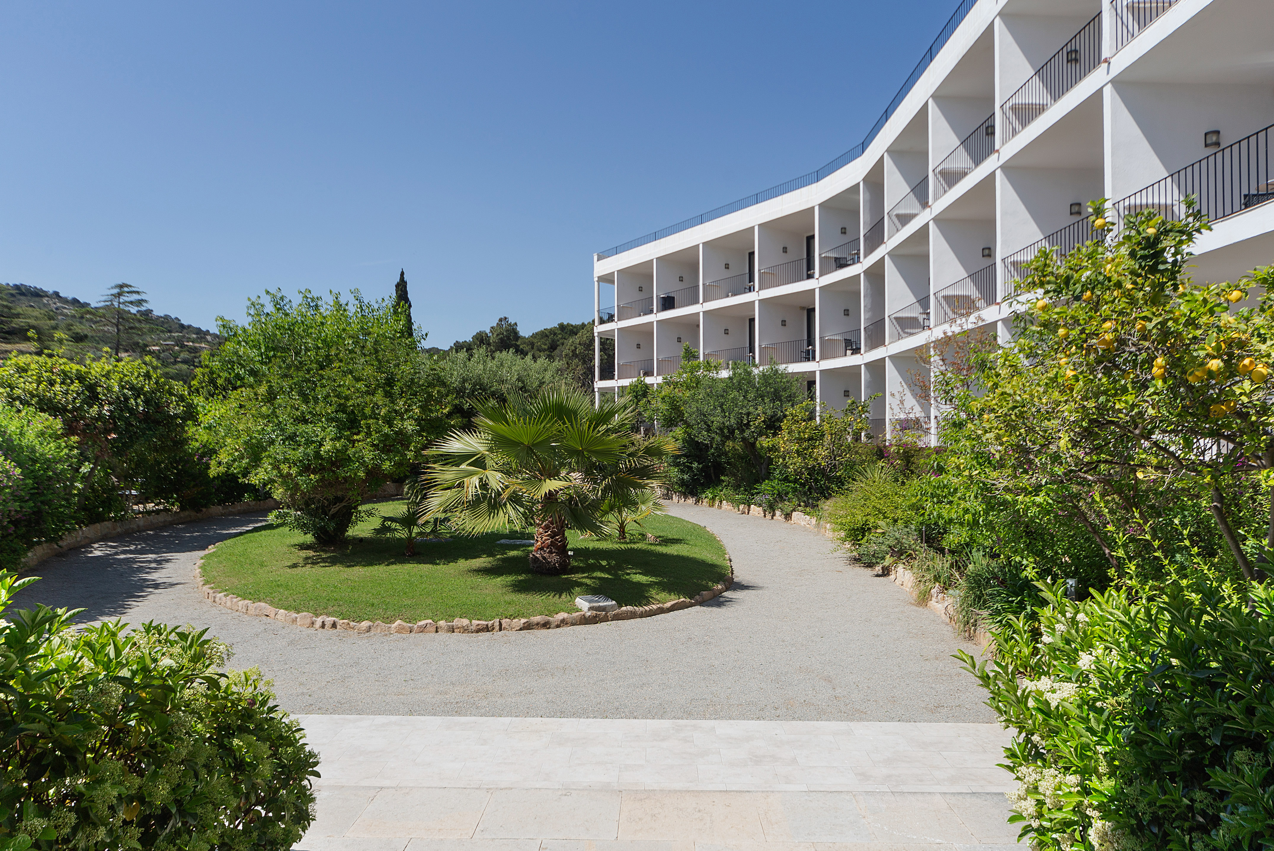 Modern curved hotel, balconies, green garden, palm trees, gravel path.