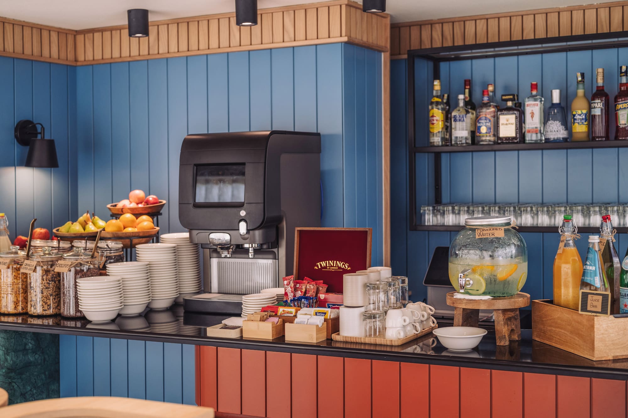Self-service breakfast counter with coffee, fresh fruit, cereals, and assorted drinks.