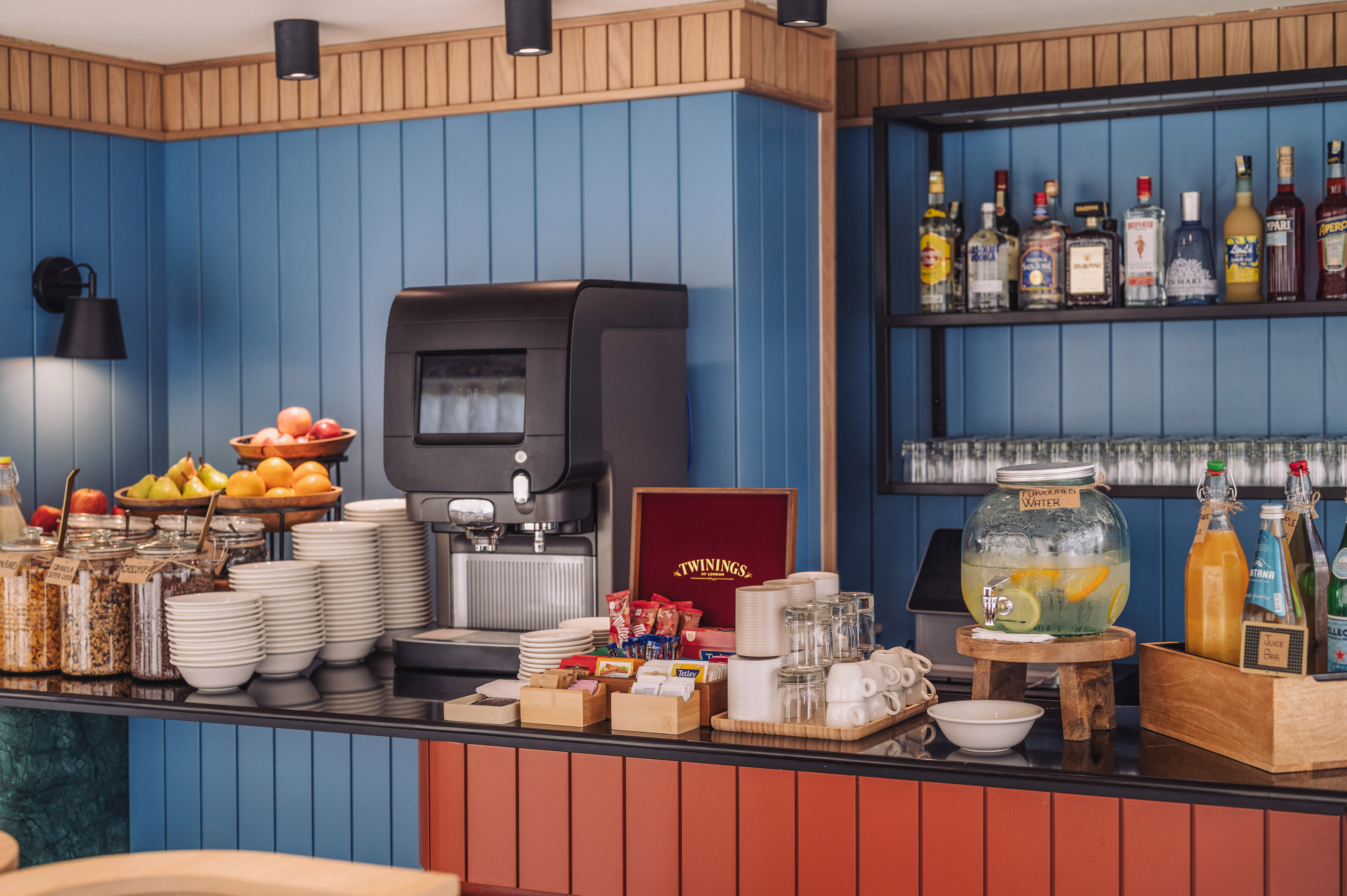 Self-service breakfast counter with coffee, fresh fruit, cereals, and assorted drinks.