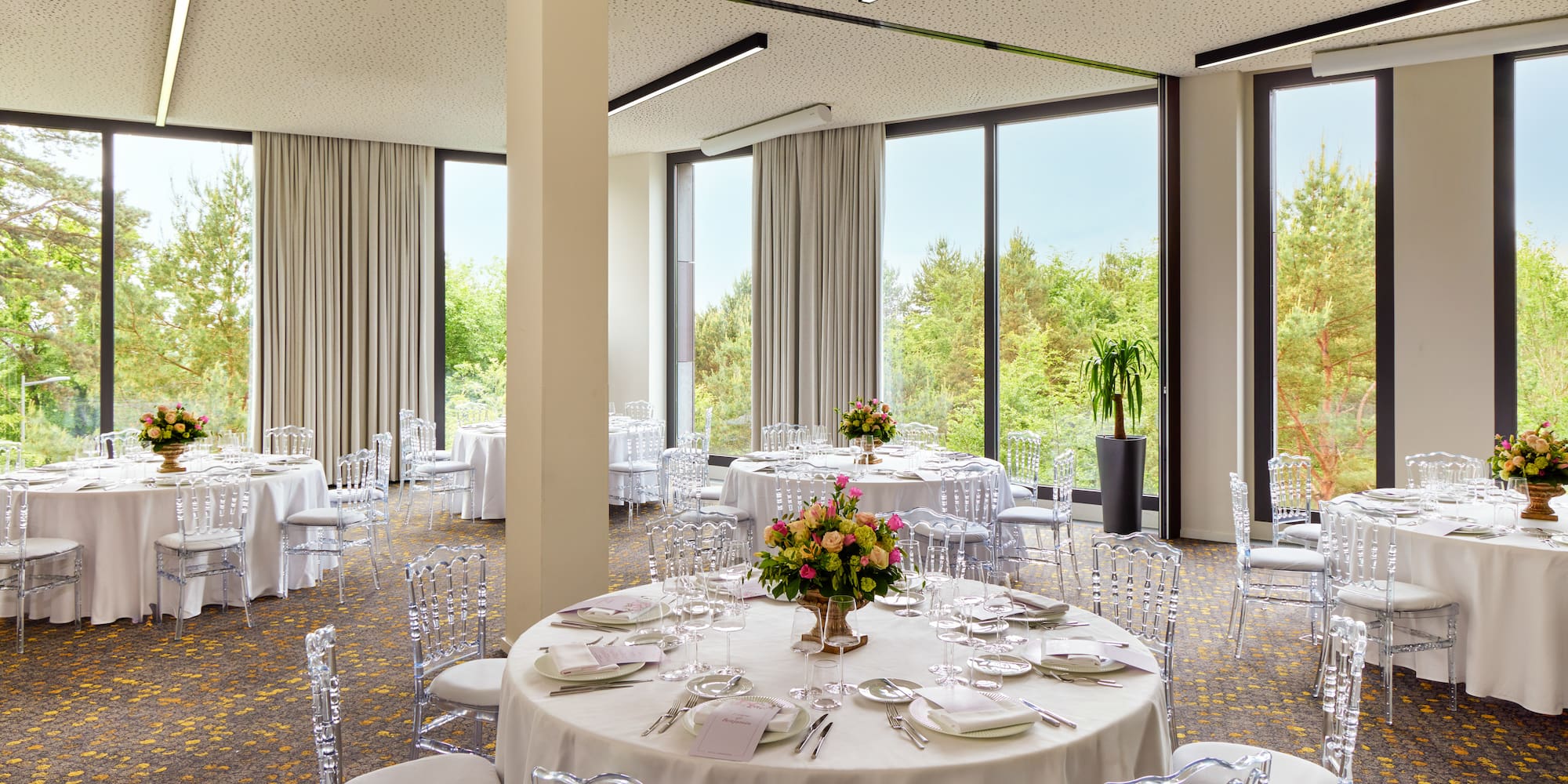Modern event room with round tables, clear chairs, flowers, and large windows.