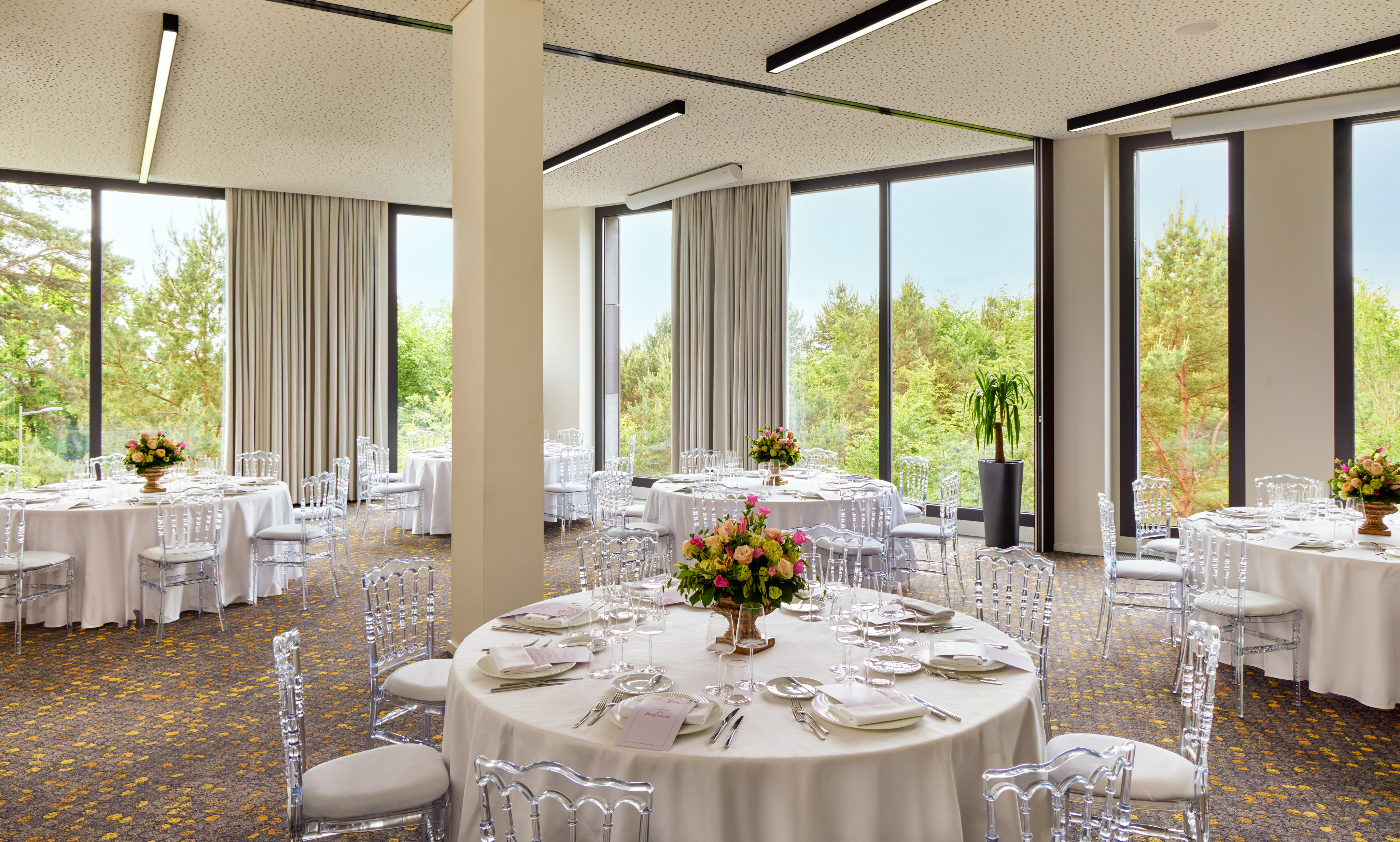 Modern event room with round tables, clear chairs, flowers, and large windows.