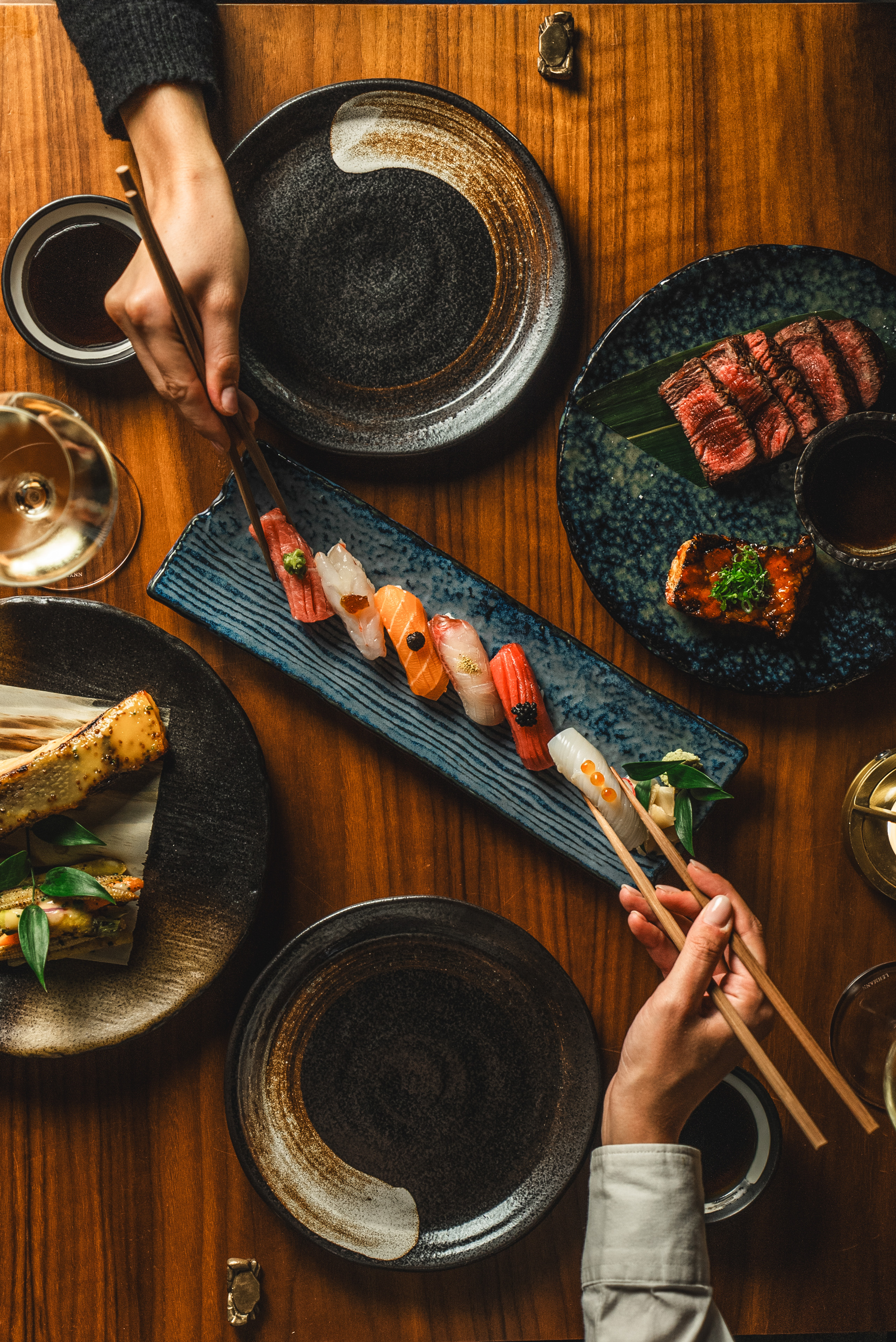 Overhead Japanese dinner: nigiri sushi, sliced beef, and hands with chopsticks.