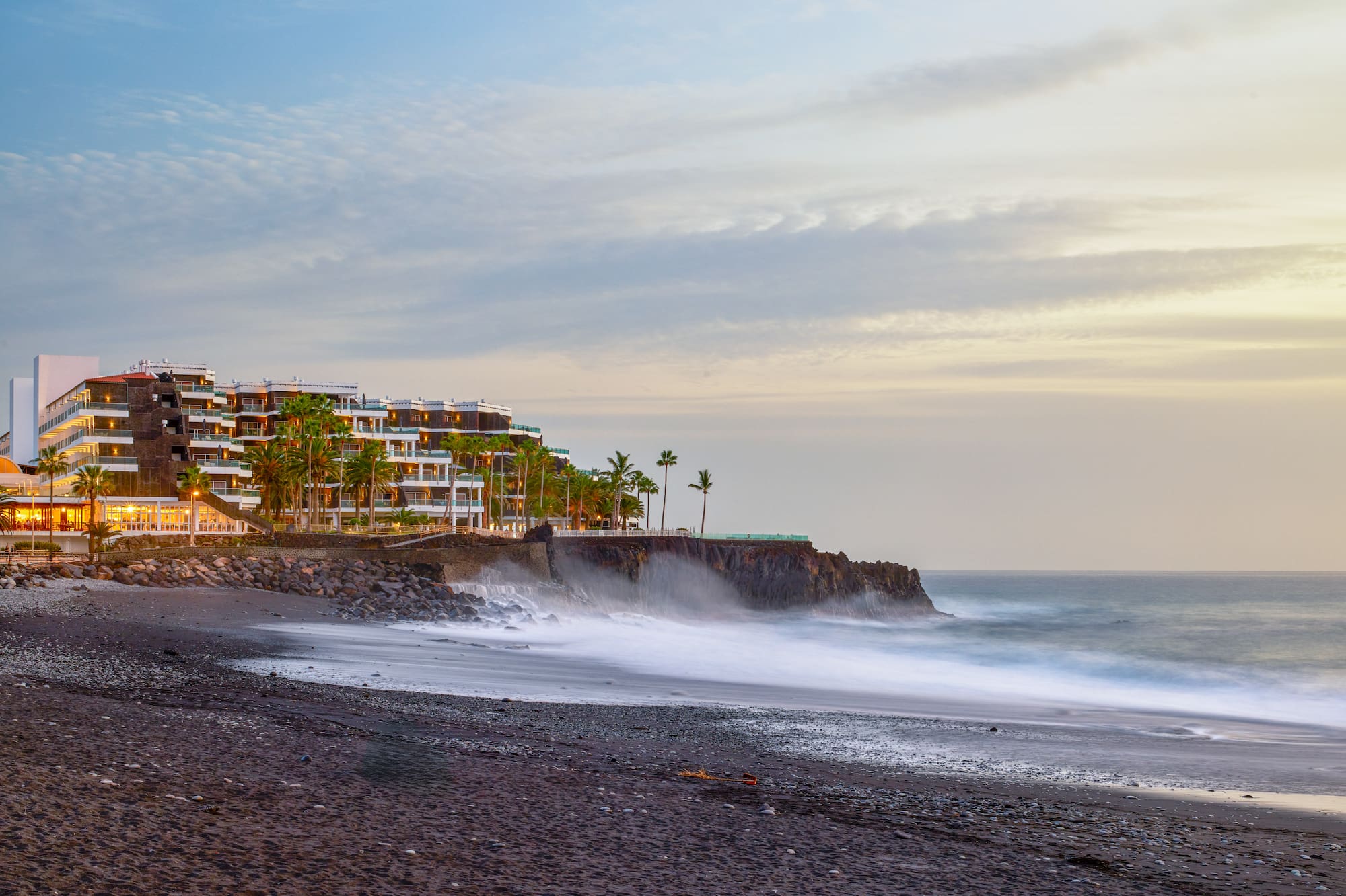 Coastal hotel on cliff, black sand beach, palm trees, and ocean waves at dusk.